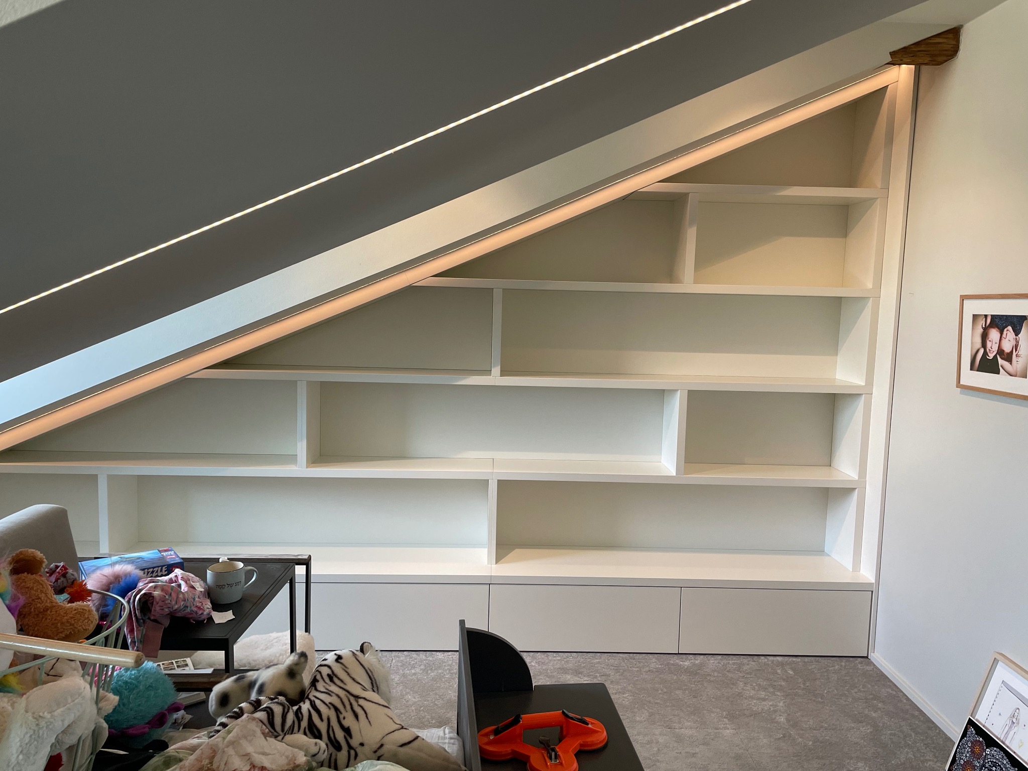 Biała, zabudowana szafa wnękowa z oświetleniem LED, dopasowana do skosu poddasza. Półki puste, w dolnej części szuflady. Ujęcie z dołu, widoczne zabawki na pierwszym planie.