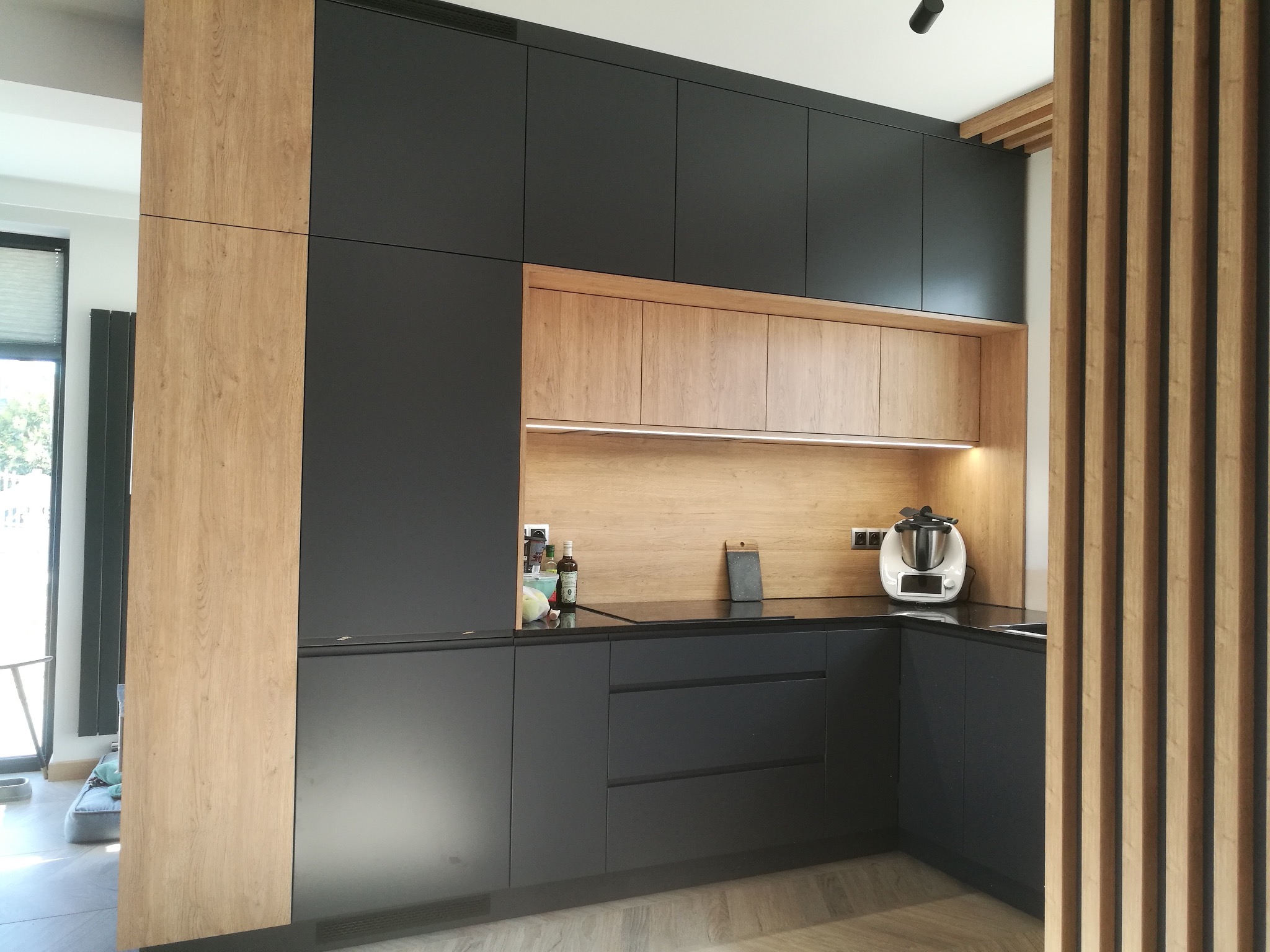 Nowoczesna kuchnia na wymiar z czarnymi frontami i drewnianymi akcentami. Widoczne szafki górne i dolne, blat oraz drewniana ścianka działowa. Oświetlenie LED pod szafkami.