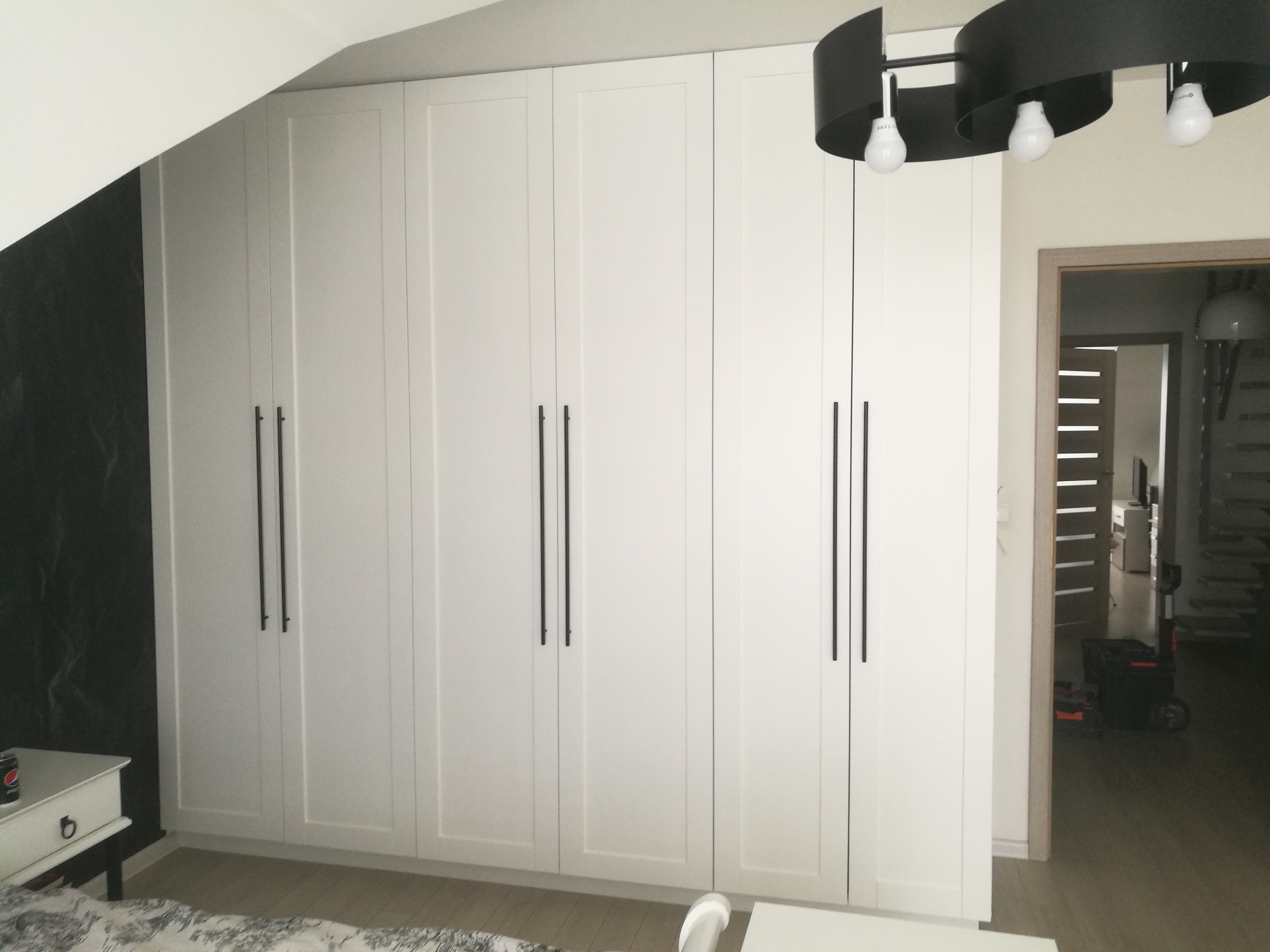 Biała szafa wnękowa z czarnymi, długimi uchwytami, dopasowana do skosu dachu, oświetlona nowoczesną lampą. W tle widoczne otwarte drzwi do innego pomieszczenia.