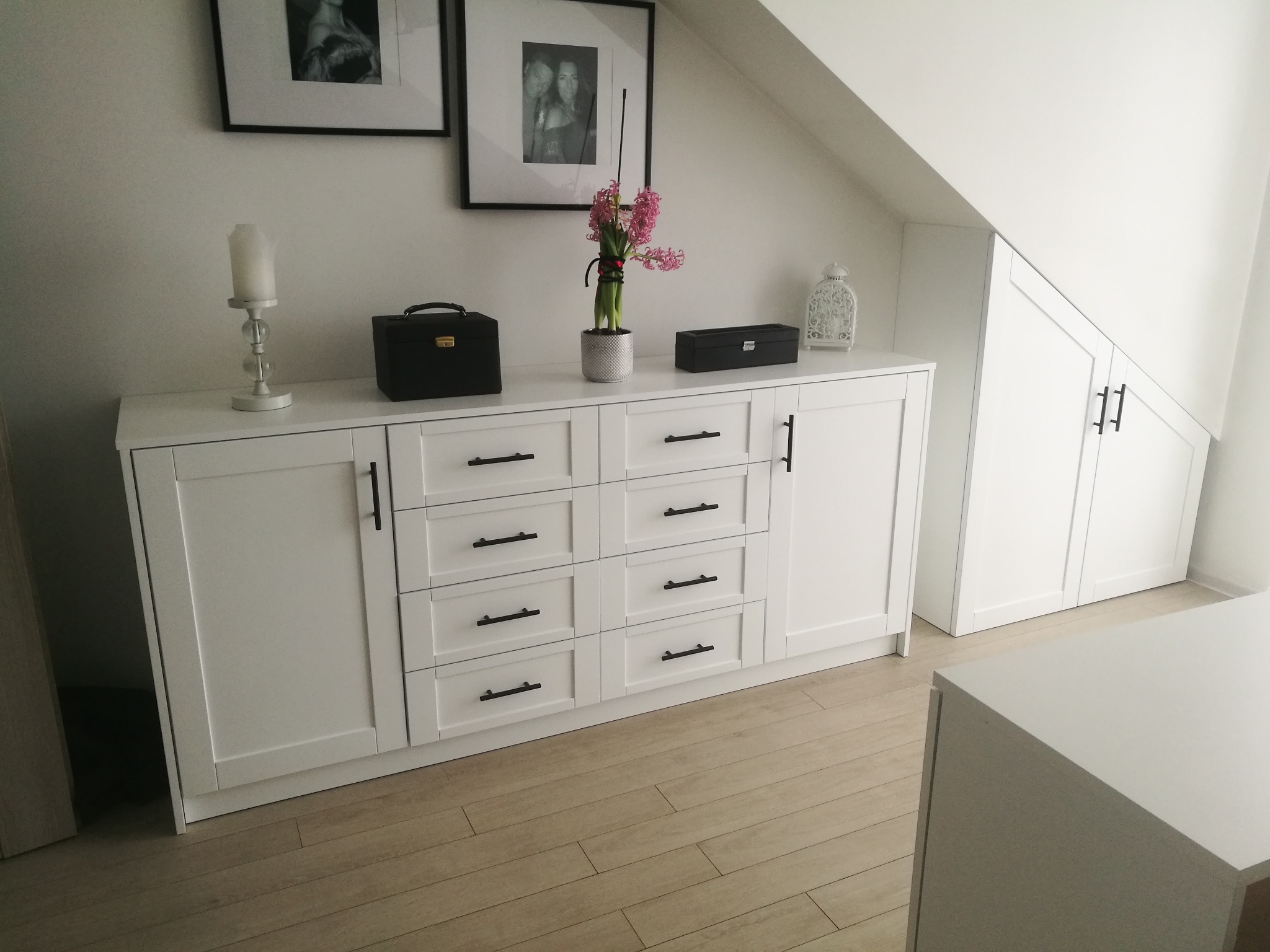 Biała komoda z szufladami i szafkami, czarne uchwyty, dekoracje na blacie, drewniana podłoga, skos dachu, minimalistyczny styl, czarno-białe zdjęcia w ramkach na ścianie.