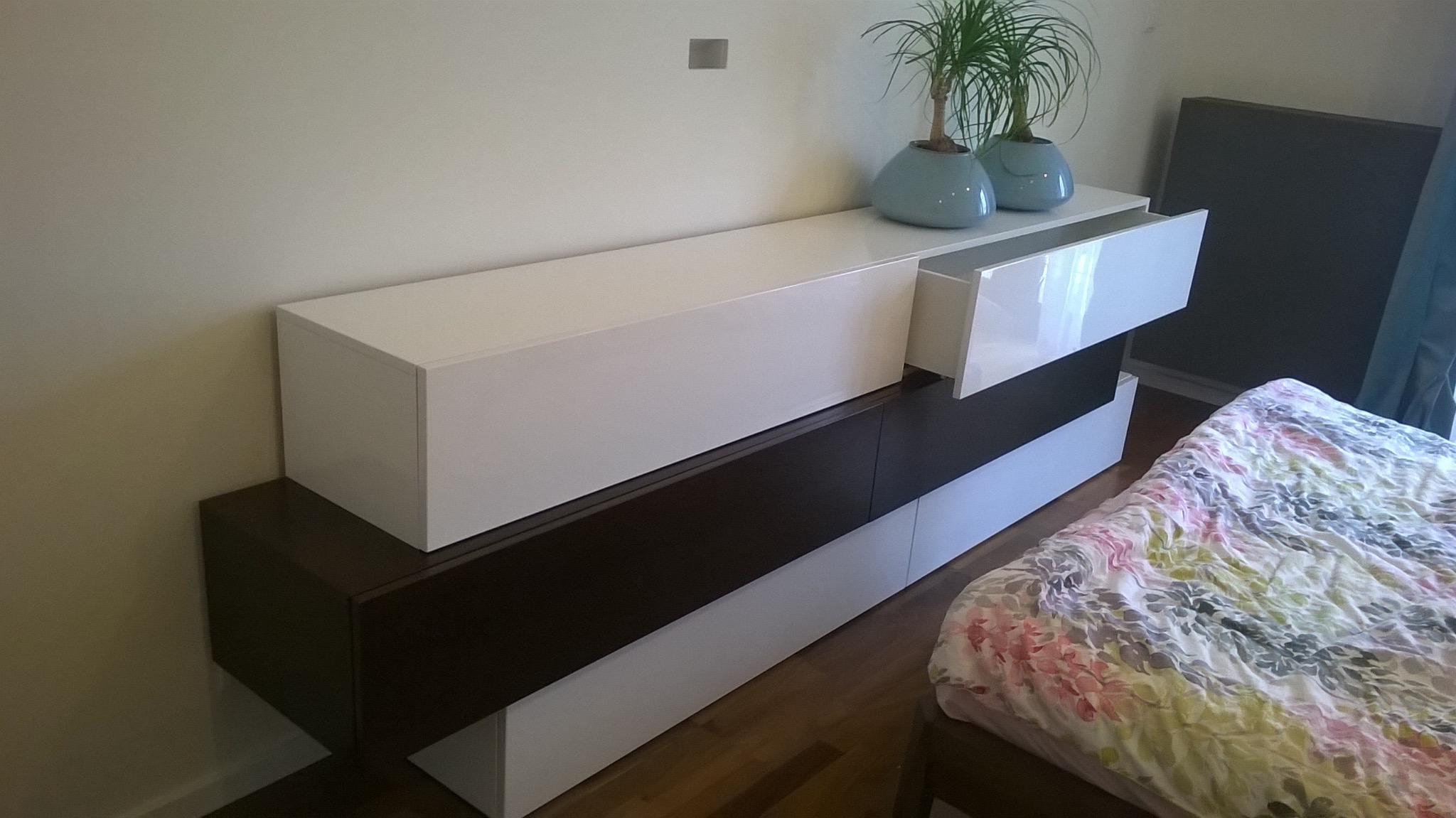 Nowoczesna szafka RTV w kolorach bieli i ciemnego brązu, z wysuwaną szufladą i dekoracyjnymi roślinami w niebieskich doniczkach na blacie, obok łóżko.