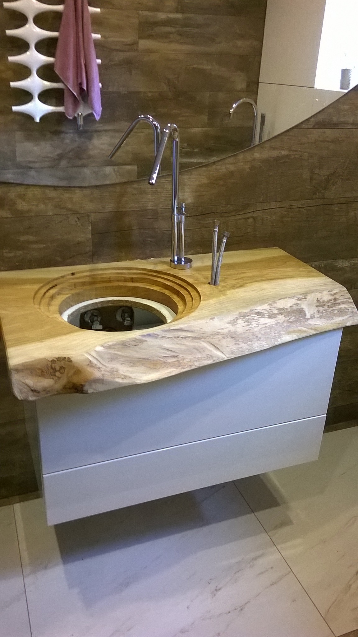 Unikatowa umywalka z blatem z litego drewna o nieregularnym kształcie, osadzona na białej szafce z szufladami. W tle brązowa ściana imitująca drewno i grzejnik.