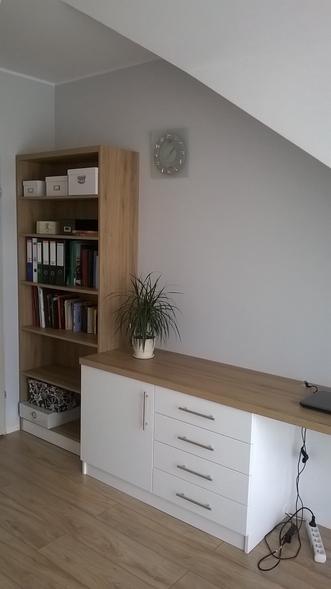 Białe szafki z drewnianym blatem i regał z książkami w pokoju na poddaszu. Na blacie doniczka z rośliną i laptop. Na ścianie zegar. Widoczne gniazdka elektryczne.