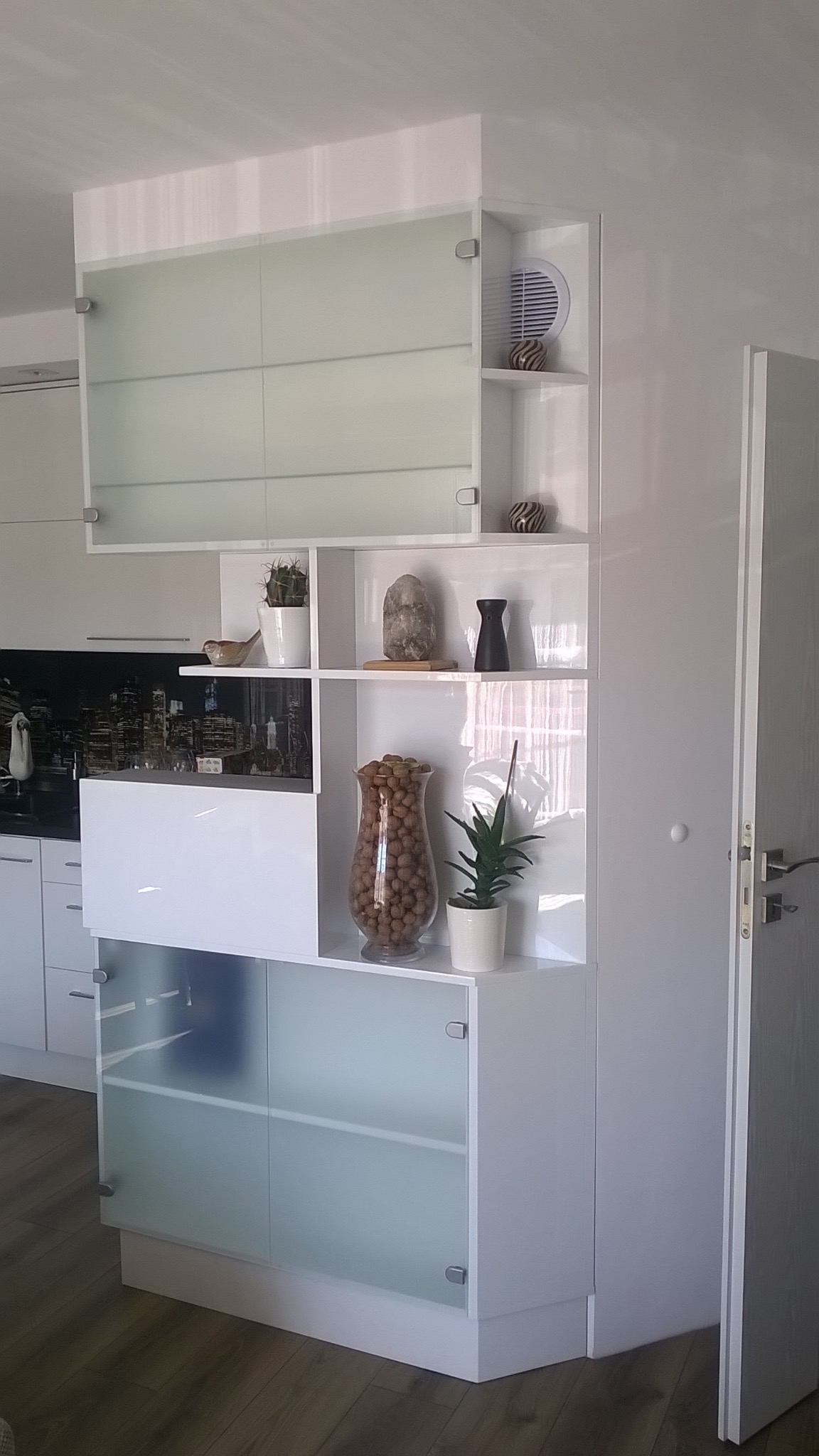 Biała, nowoczesna szafa wnękowa z półkami i przeszklonymi drzwiczkami, dekorowana elementami wystroju wnętrz. Widoczny fragment kuchni w tle.