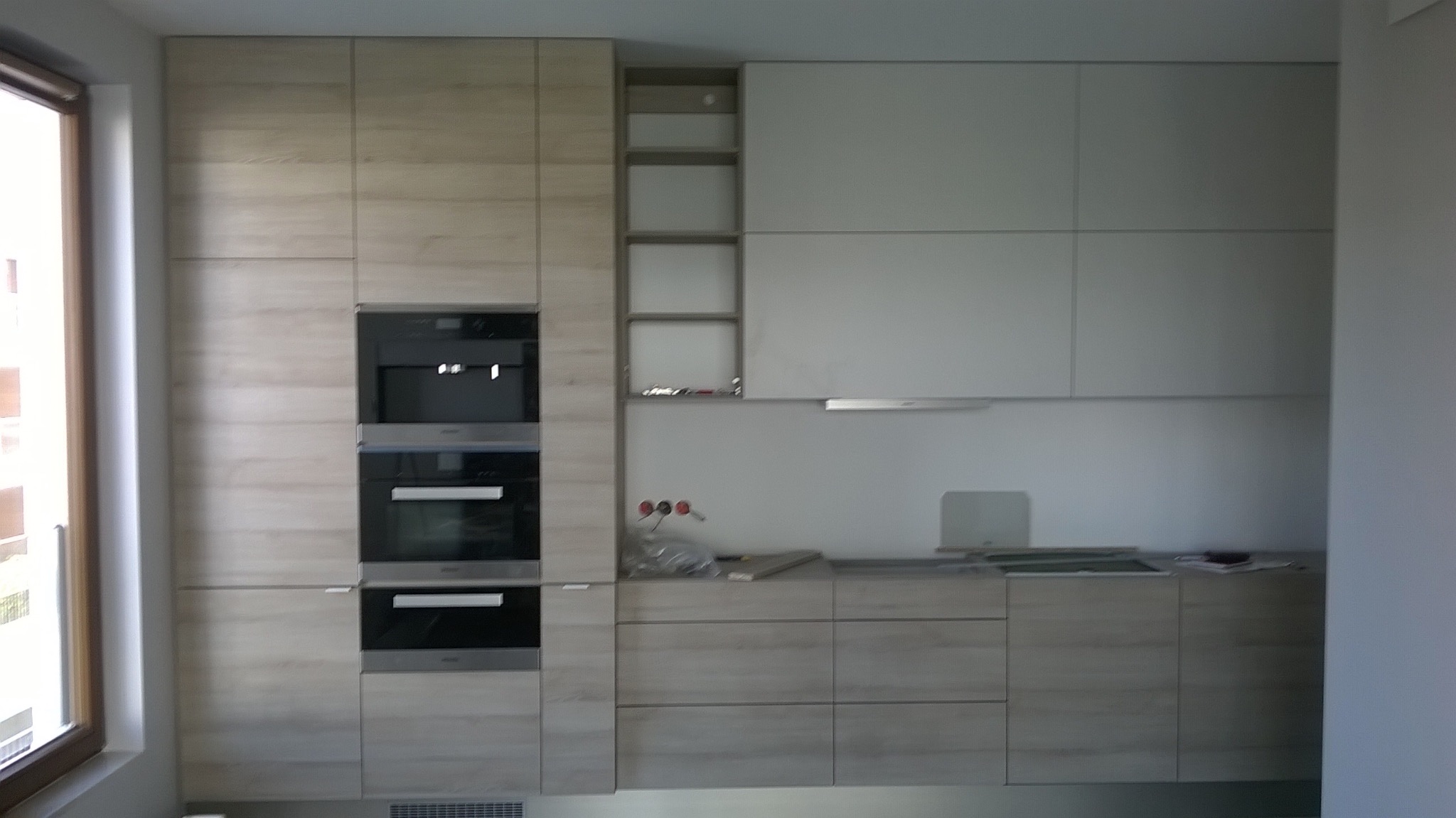 Jasna kuchnia z drewnianymi frontami i wbudowanym sprzętem AGD, minimalistyczny design, oświetlenie podszafkowe, widok na okno z lewej strony.