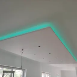 Podwieszany sufit z oświetleniem LED w kolorze turkusowym, widoczne otwory na punkty świetlne, surowe ściany w trakcie wykańczania.