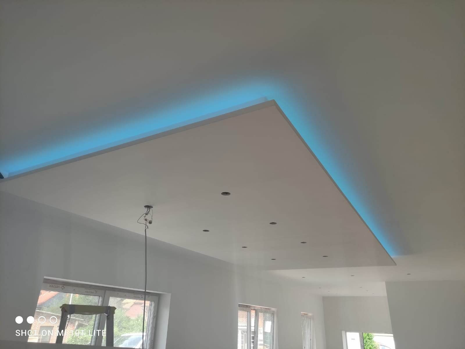 Wykończony sufit podwieszany z oświetleniem LED w kolorze błękitnym, widoczne otwory na oświetlenie punktowe, surowe ściany, okna i drabina w tle.