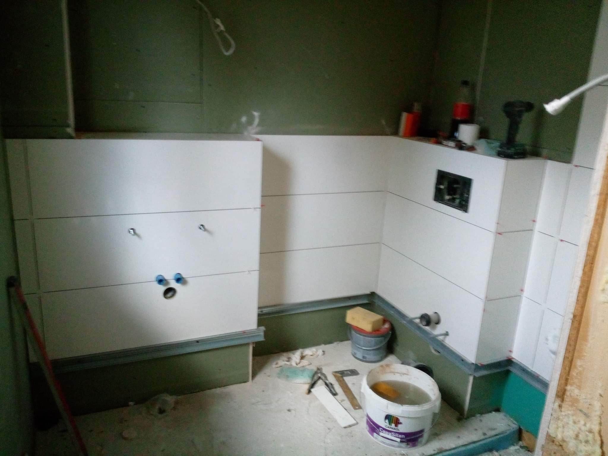 Białe płytki ułożone na ścianach w trakcie prac w łazience, widoczne narzędzia, wiadro z wodą i gąbka, ściany częściowo wykończone płytami gipsowo-kartonowymi.