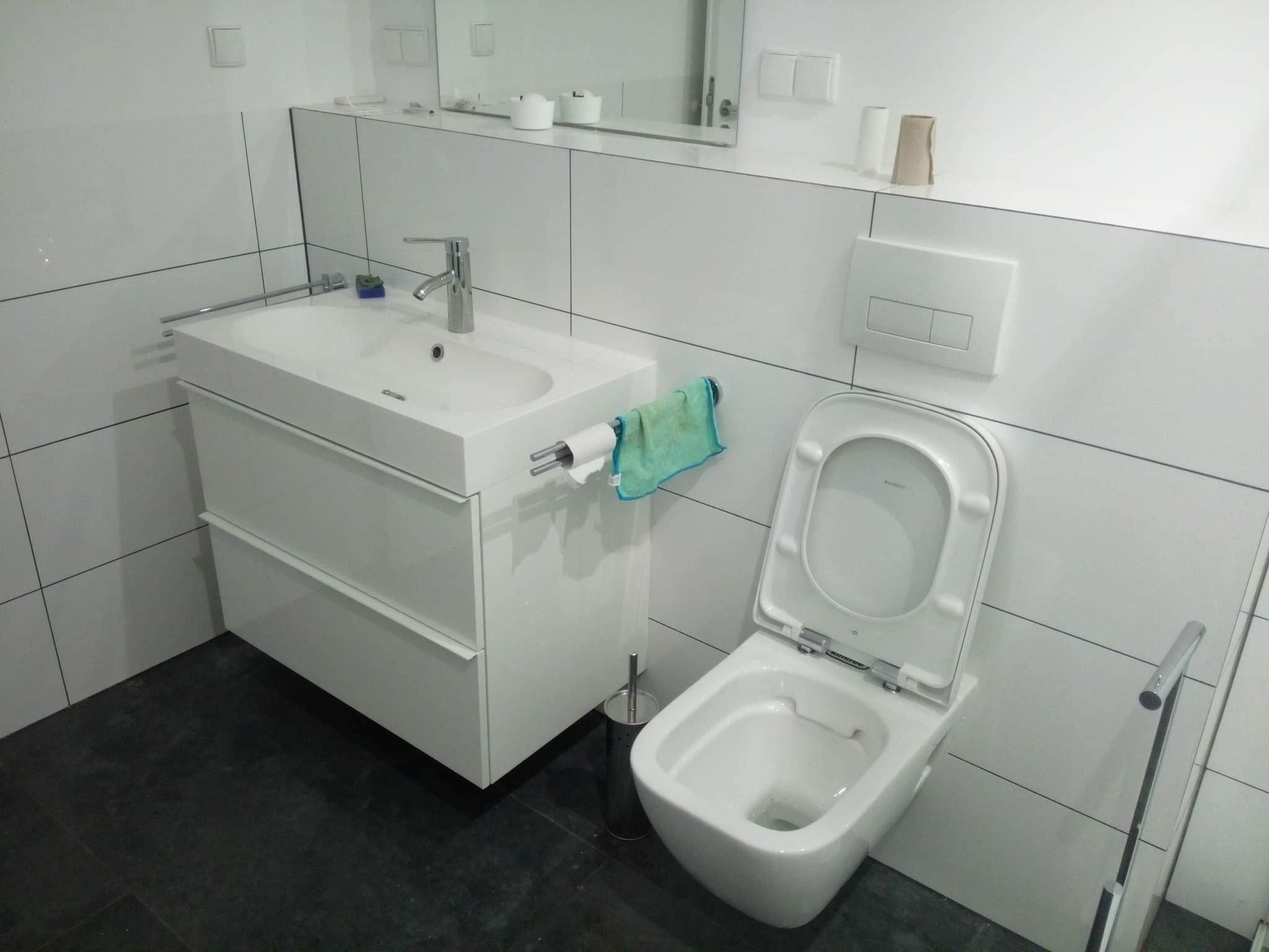 Nowoczesna łazienka z białą armaturą i meblami, czarne płytki podłogowe, minimalistyczny design, toaleta z deską wolnoopadającą, umywalka z szafką i chromowaną baterią, zielony ręcznik.