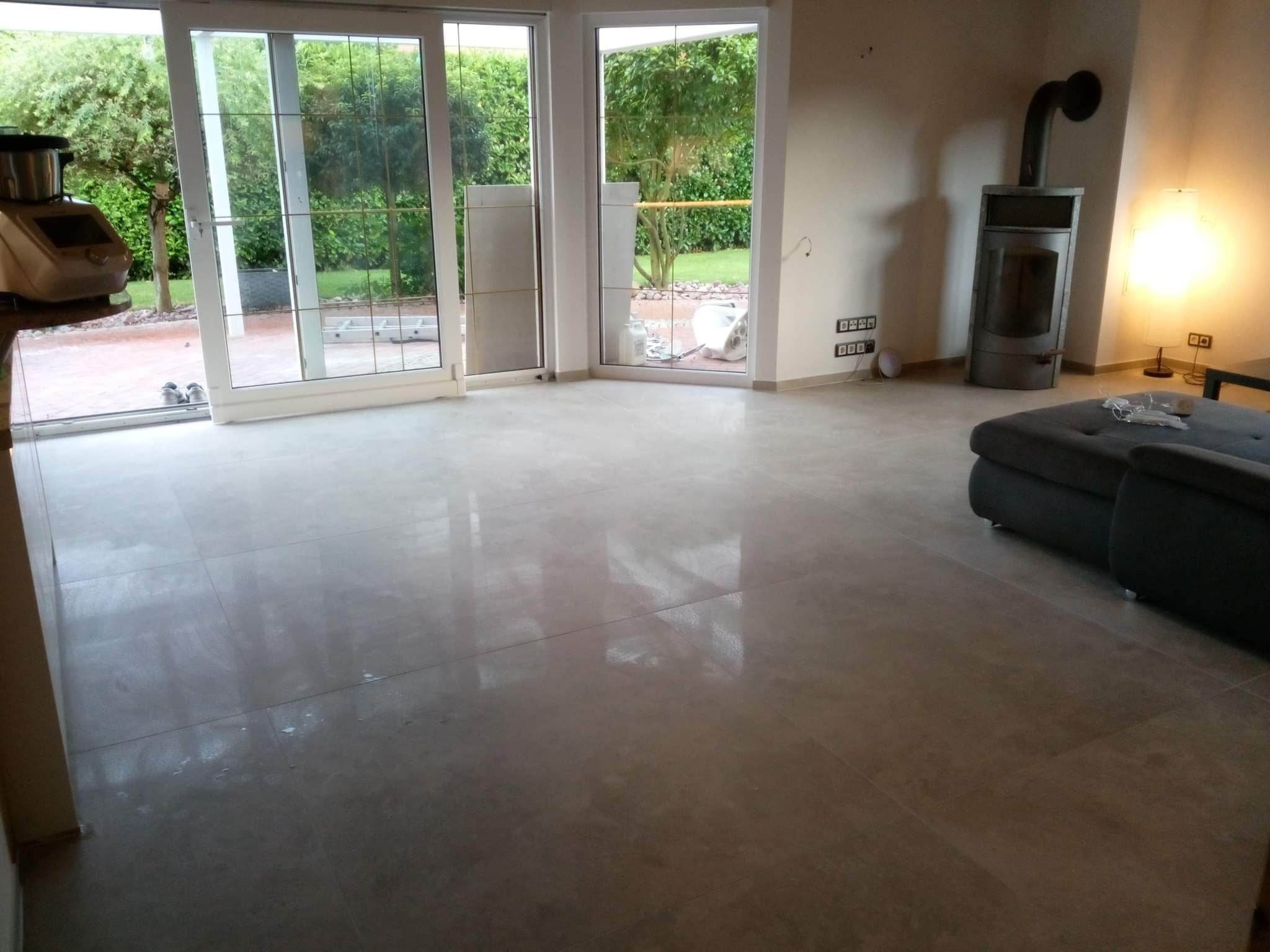 Wnętrze domu po remoncie z nową, połyskującą podłogą z płytek, widoczne otwarte okno tarasowe z widokiem na ogród, piec wolnostojący i fragment szarej sofy.