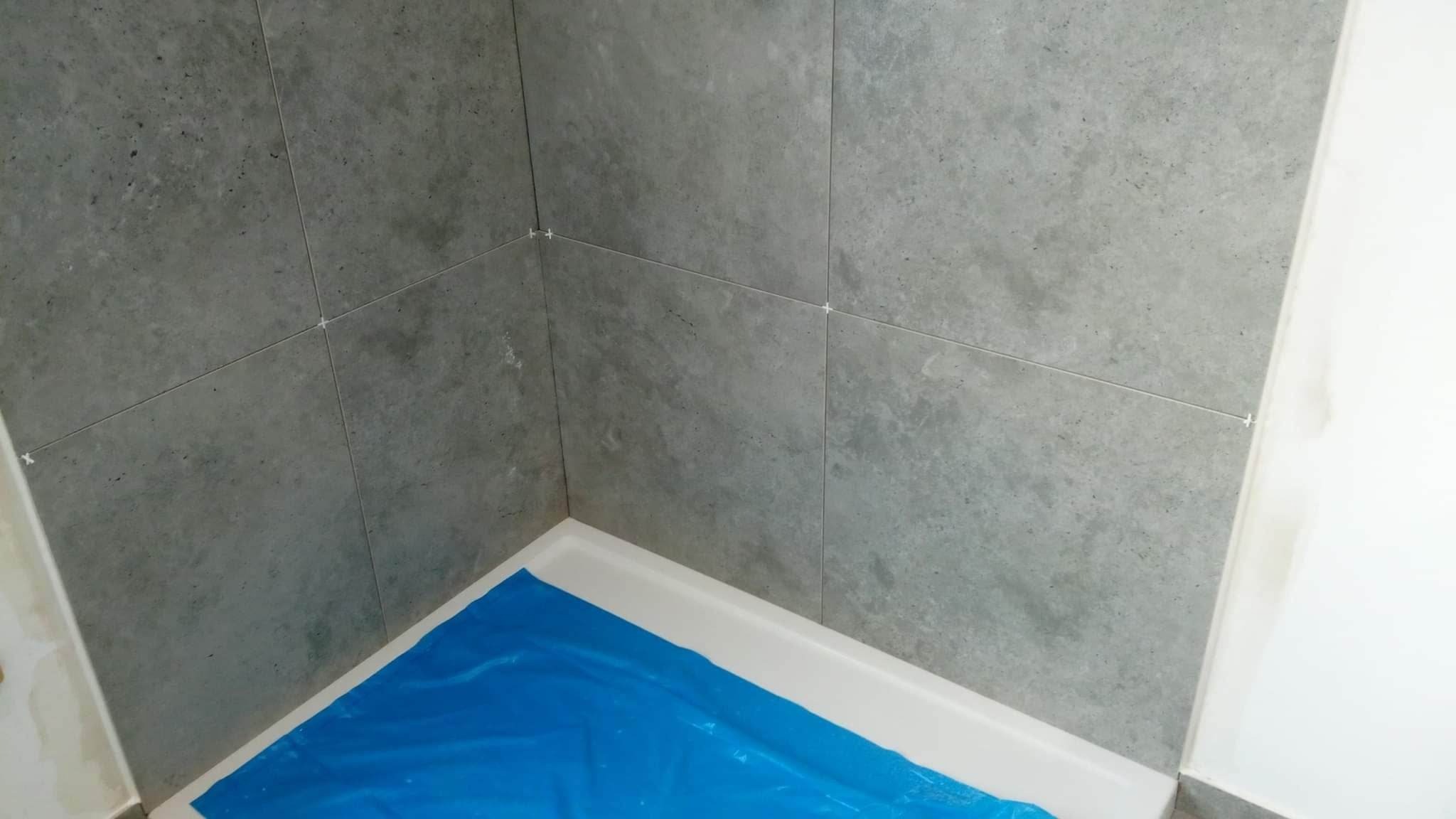 Świeżo ułożone, szare płytki w narożniku kabiny prysznicowej, z widocznymi krzyżykami dystansowymi i zabezpieczoną folią brodzikiem.