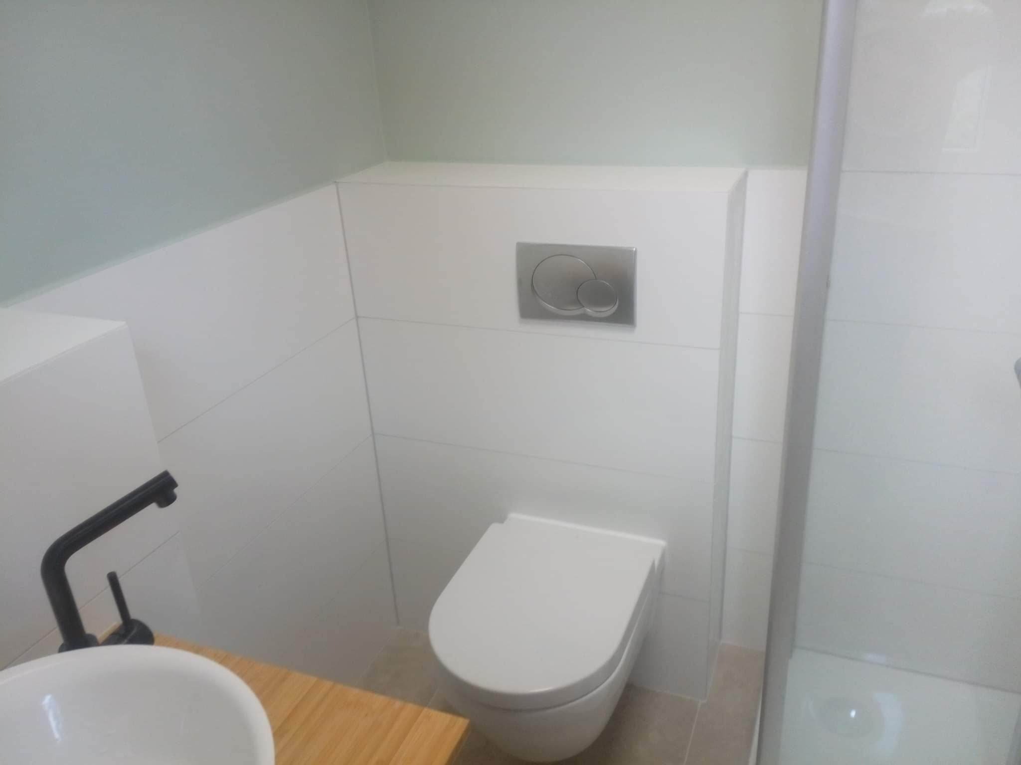 Nowoczesna łazienka z podwieszaną toaletą, chromowanym przyciskiem spłukującym, umywalką nablatową z czarną armaturą i białymi płytkami ściennymi.