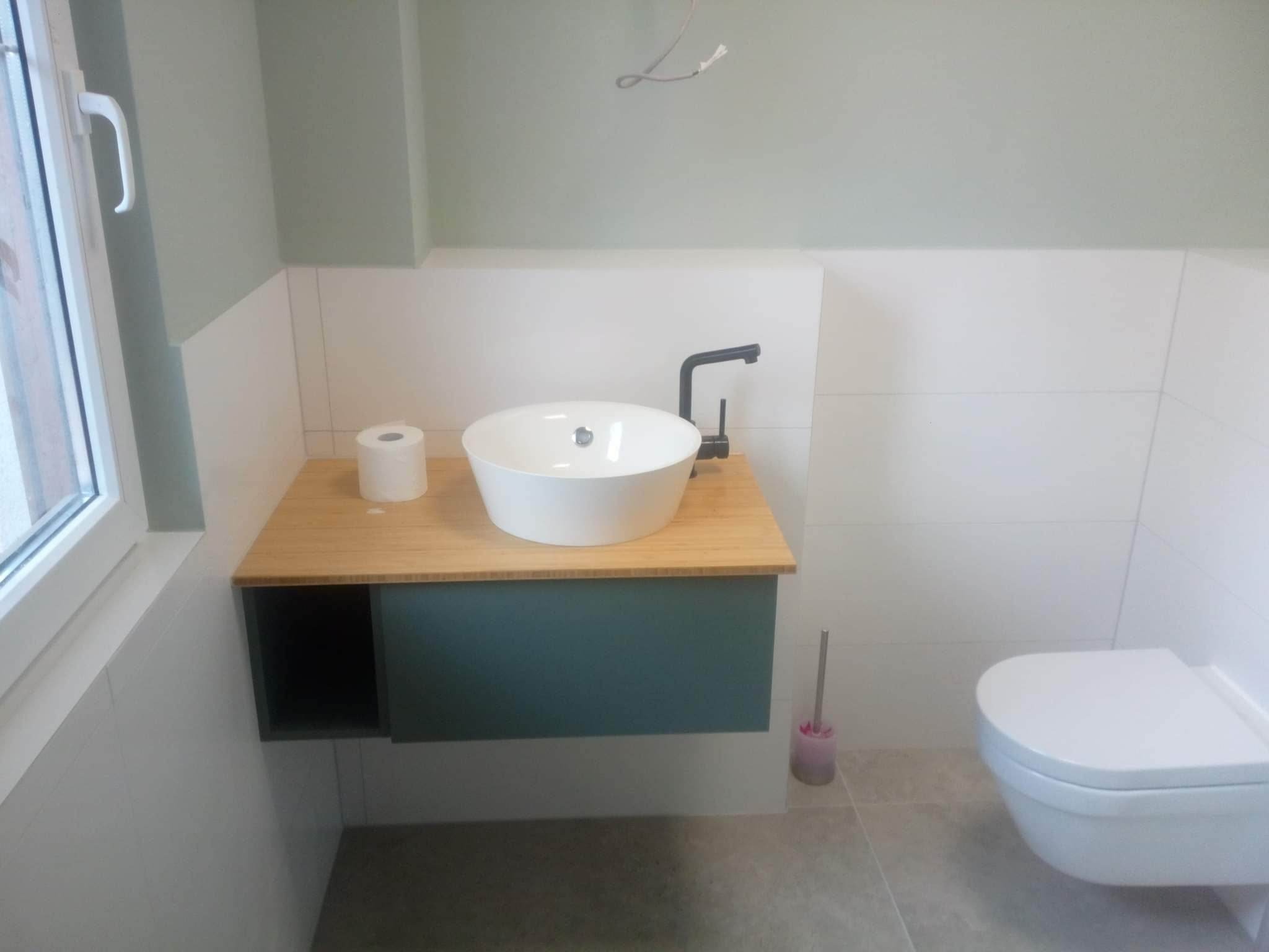 Nowoczesna łazienka z wiszącą toaletą, okrągłą umywalką nablatową z czarną armaturą, drewnianym blatem i zielonymi akcentami kolorystycznymi na tle białych płytek.