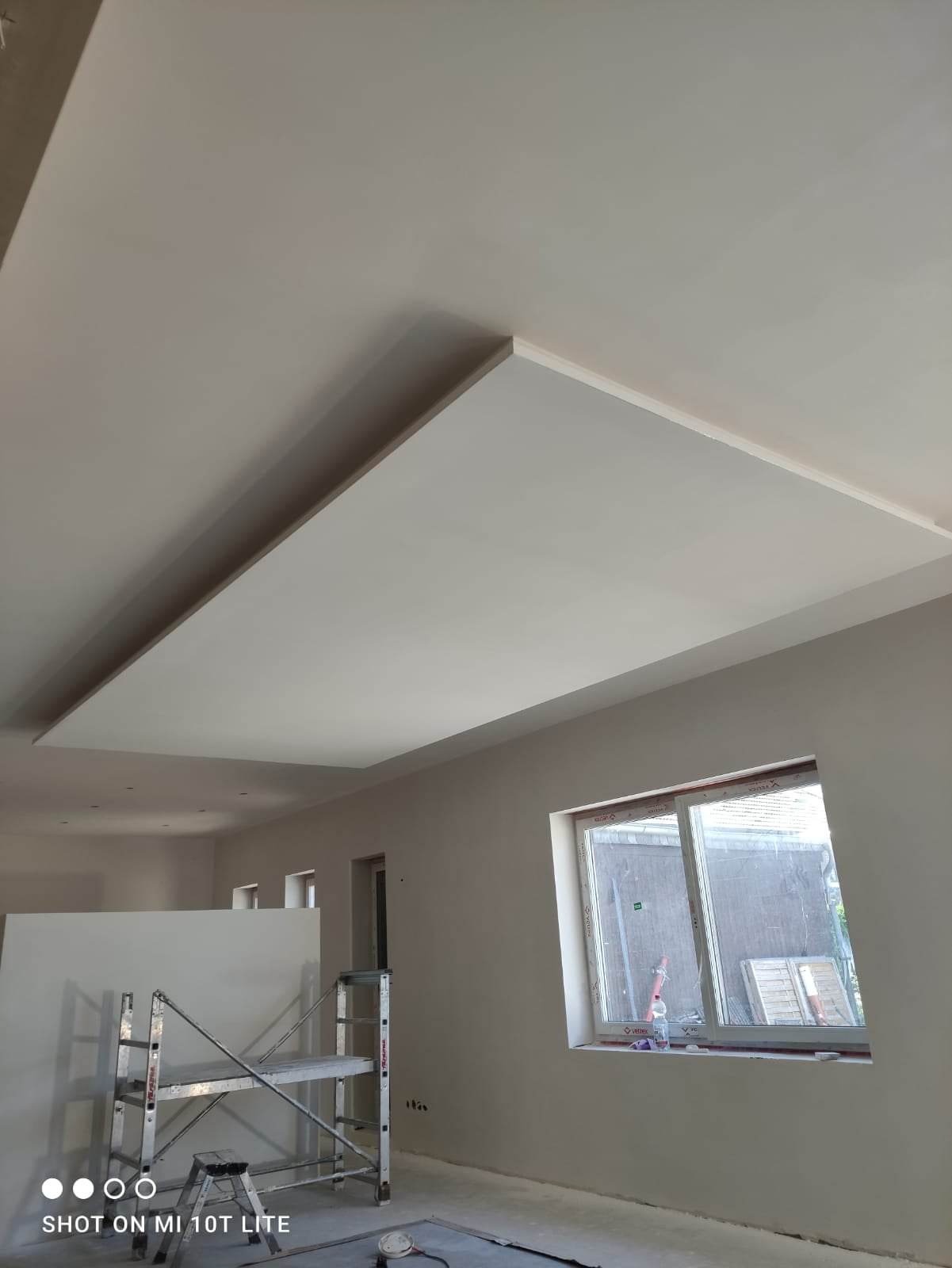 Wykończone wnętrze z podwieszanym sufitem oświetlonym wnęką, widoczne rusztowanie i okno z widokiem na zewnątrz.