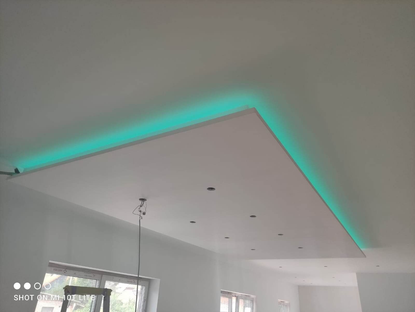 Podwieszany sufit z oświetleniem LED w kolorze turkusowym, widoczne otwory na punkty świetlne, surowe ściany w trakcie wykańczania.