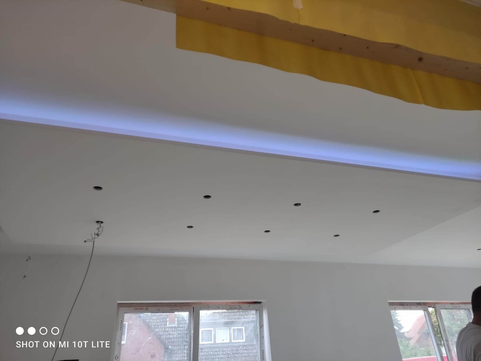 Biały sufit podwieszany z oświetleniem LED i otworami na punkty świetlne, widoczne okna z białymi ramami i fragment ocieplenia poddasza z żółtą folią.