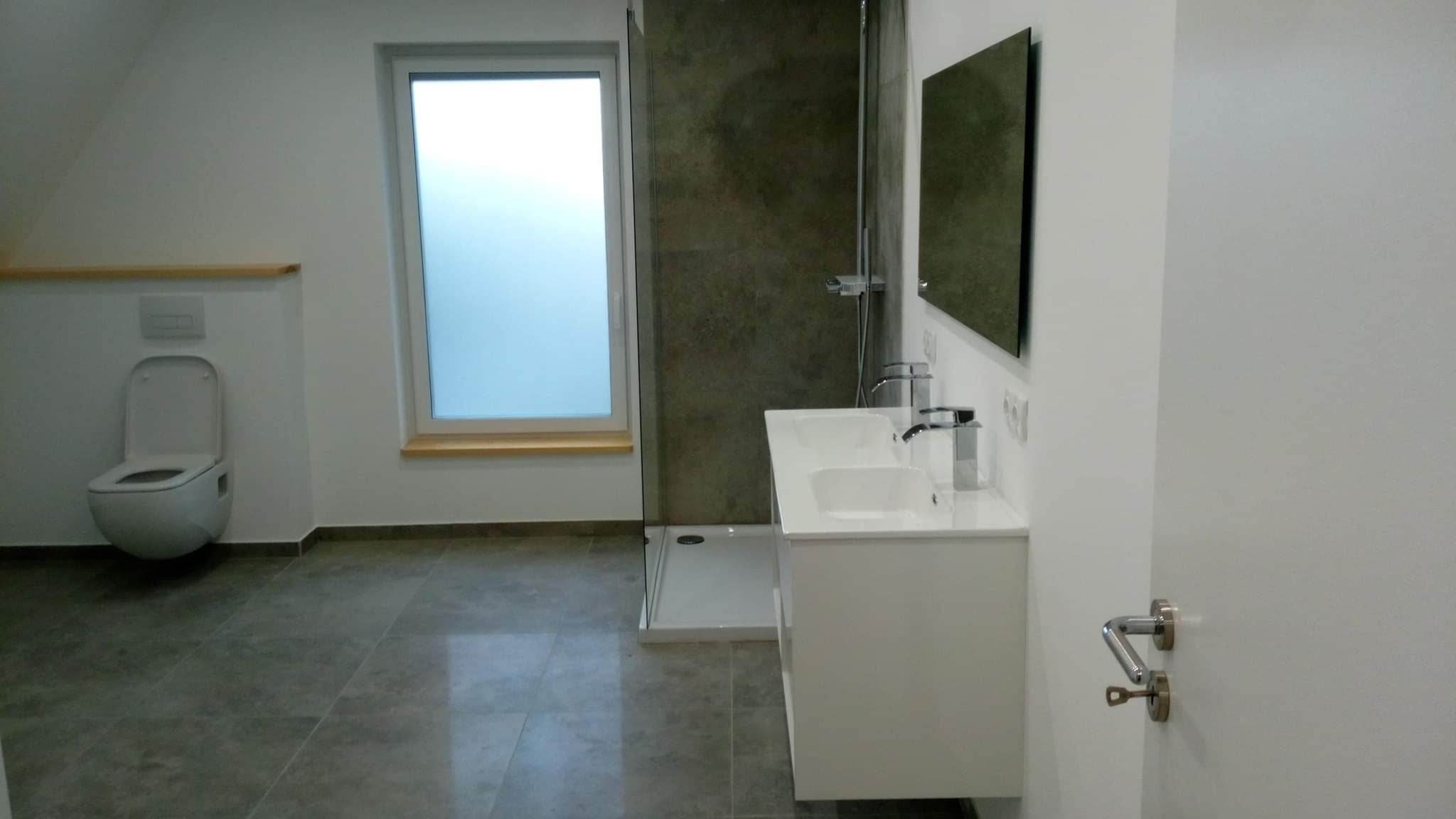 Nowoczesna łazienka z podwójną umywalką, kabiną prysznicową z szarymi płytkami imitującymi beton, wiszącą toaletą i dużym oknem z matową szybą.