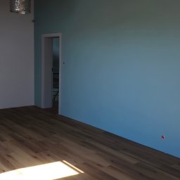 Alex Oleksandr Lisovyi - Pomalowane na niebiesko ściany pokoju z nową drewnianą podłogą, widoczny fragment oświetlenia sufitowego i otwarte drzwi do drugiego pokoju.