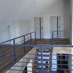 Alex Oleksandr Lisovyi - Wnętrze domu w trakcie wykańczania: drewnopodobna podłoga, schody z szarymi nakładkami, metalowa barierka i białe ściany z dwojgiem drzwi.