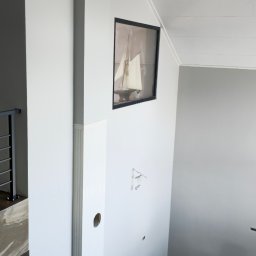 Alex Oleksandr Lisovyi - Wnętrze domu w trakcie wykańczania: białe ściany, drewniana podłoga, widoczne niedokończone instalacje elektryczne, obraz z motywem żaglówki na ścianie, narożnik i fotel w jasnej kolorystyce.