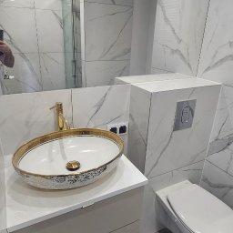 Alex Oleksandr Lisovyi - Elegancka łazienka po remoncie z owalną umywalką w kolorze białym ze złotymi zdobieniami, złotą baterią, podwieszaną toaletą i płytkami imitującymi marmur na ścianach.