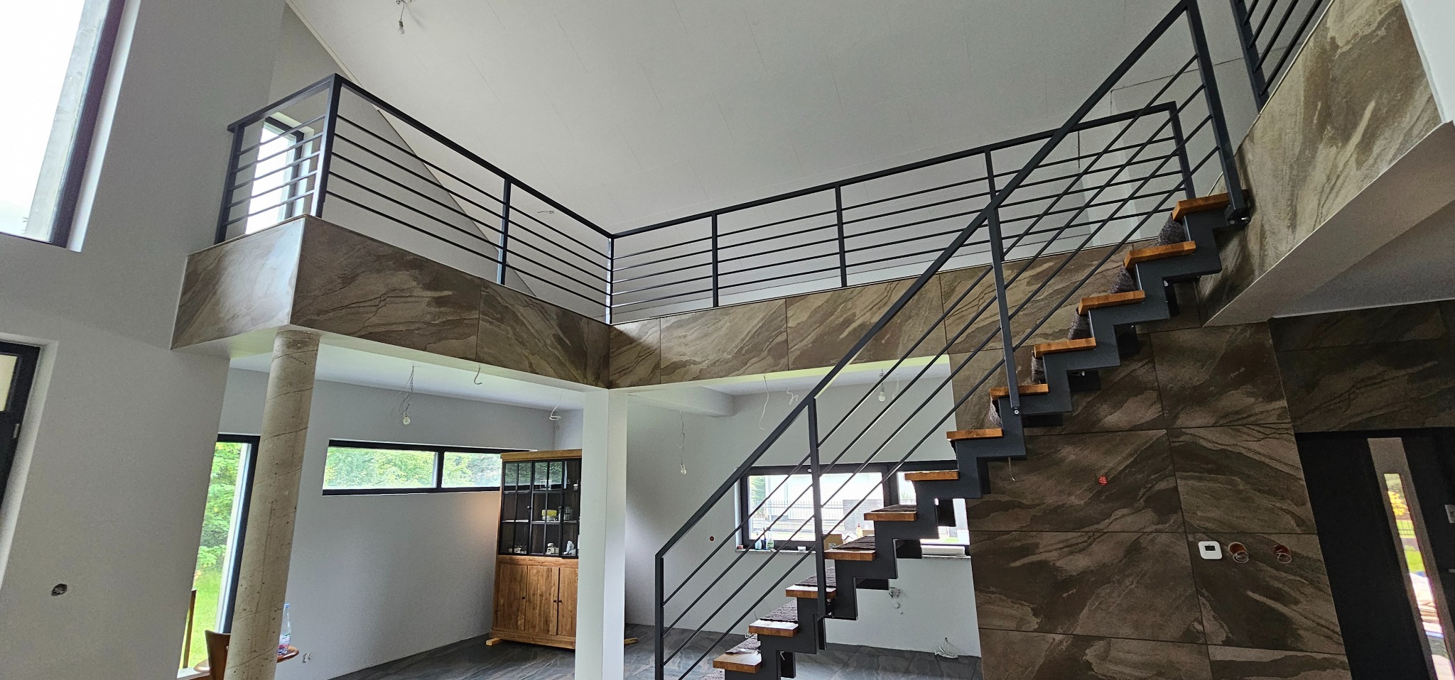 Dwupoziomowe wnętrze z czarnymi schodami z drewnianymi stopniami i marmurową ścianą, widok z dołu na antresolę z czarną balustradą i marmurowym wykończeniem, w tle przeszklona witryna.