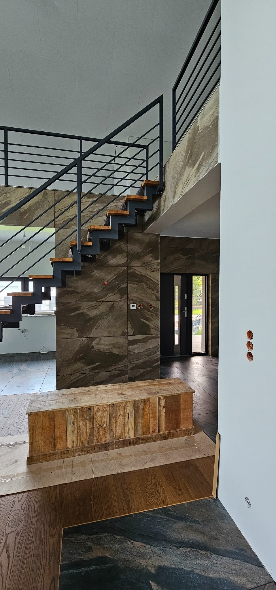 Wnętrze domu w trakcie prac wykończeniowych: widoczne schody z metalową balustradą i drewnianymi stopniami, ściany wyłożone płytkami imitującymi kamień, drewniana komoda na podłodze, częściowo...