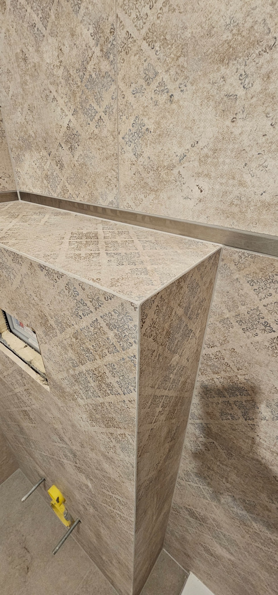 Obudowa stelaża podtynkowego WC wykończona beżowymi płytkami z delikatnym, ciemniejszym wzorem kwiatowym, widoczne wystające elementy montażowe.