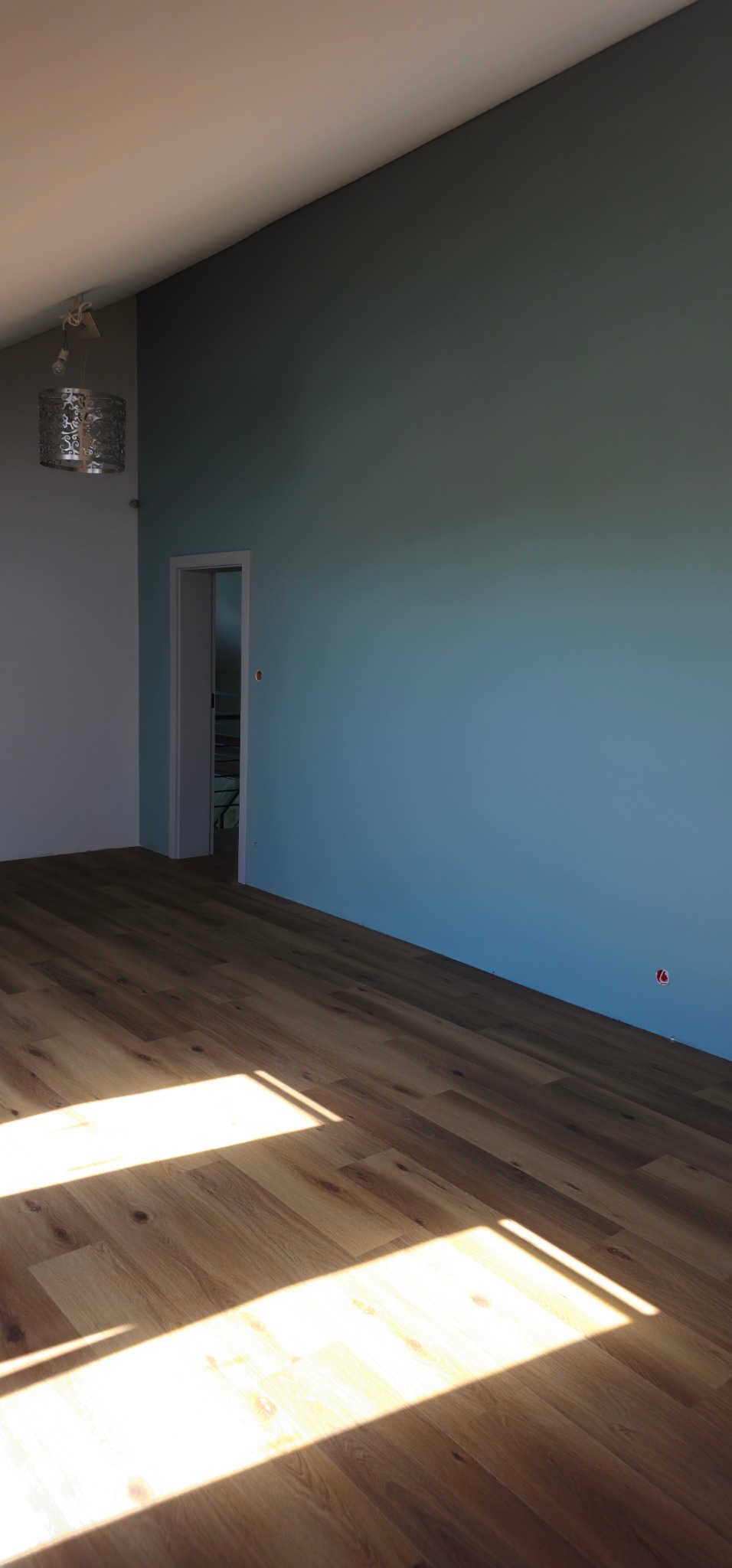 Pomalowane na niebiesko ściany pokoju z nową drewnianą podłogą, widoczny fragment oświetlenia sufitowego i otwarte drzwi do drugiego pokoju.