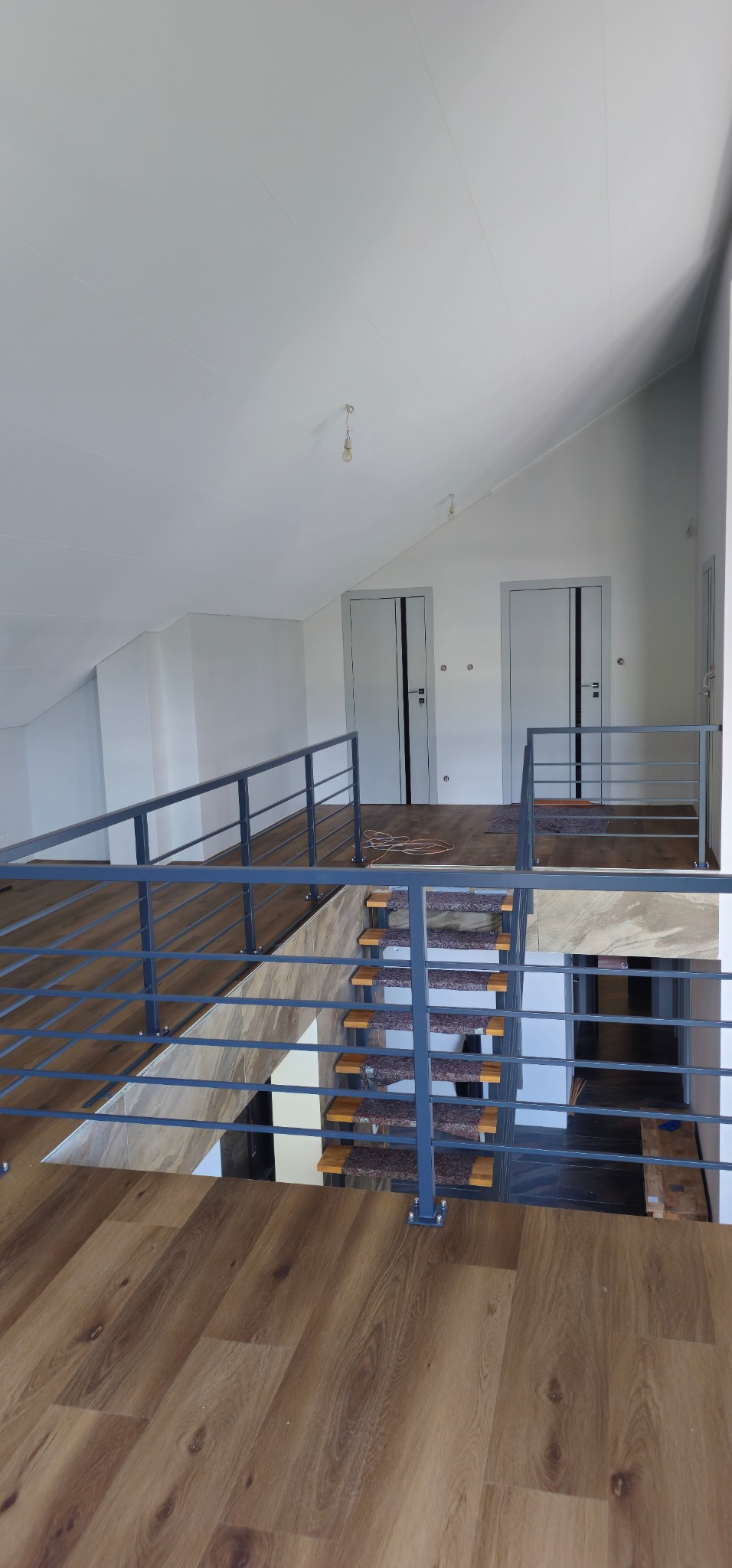 Wnętrze domu w trakcie wykańczania: drewnopodobna podłoga, schody z szarymi nakładkami, metalowa barierka i białe ściany z dwojgiem drzwi.