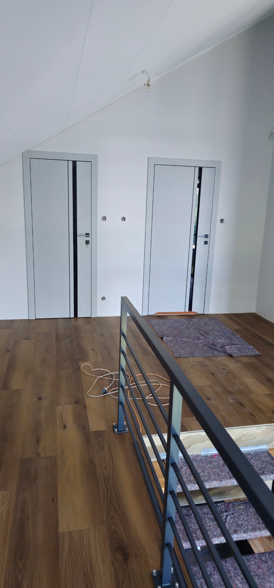 Nowoczesne, białe drzwi z czarnymi akcentami, zamontowane w jasnym pomieszczeniu z drewnianą podłogą, widoczne z perspektywy schodów z metalową barierką.