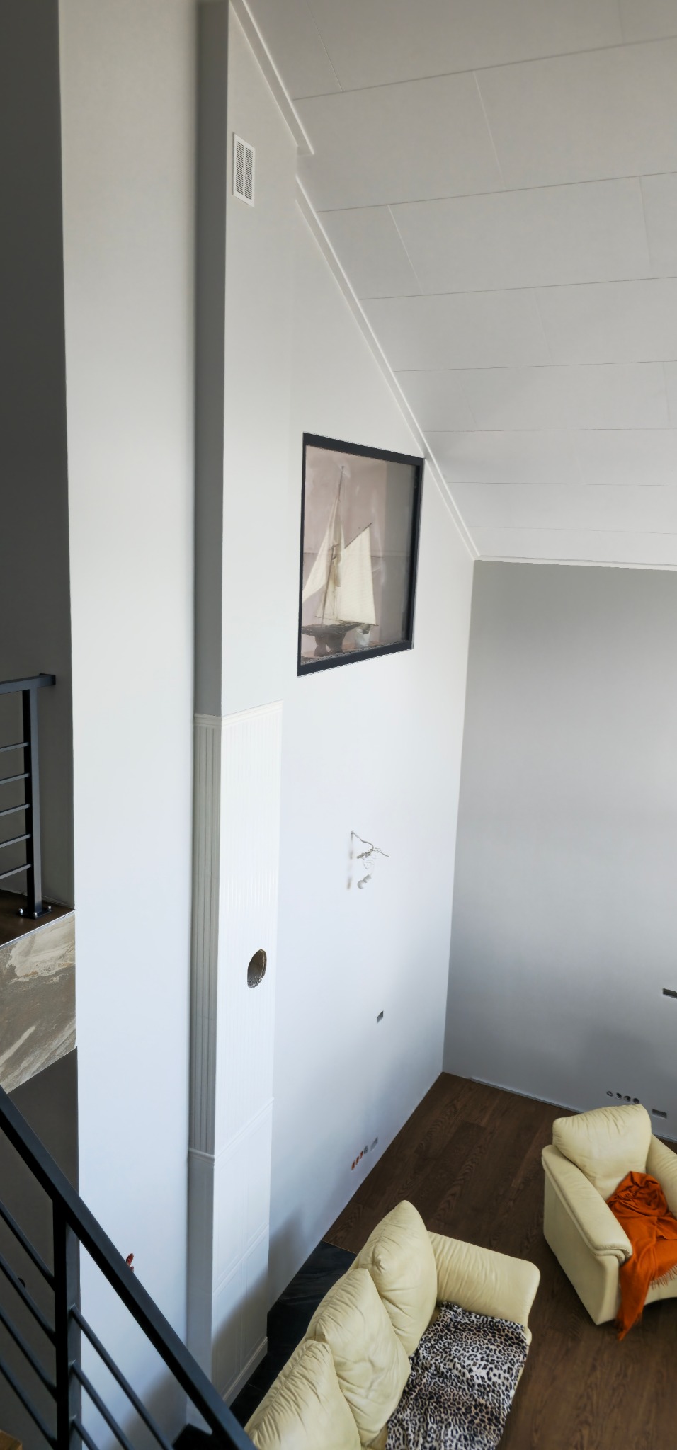 Wnętrze domu w trakcie wykańczania: białe ściany, drewniana podłoga, widoczne niedokończone instalacje elektryczne, obraz z motywem żaglówki na ścianie, narożnik i fotel w jasnej kolorystyce.