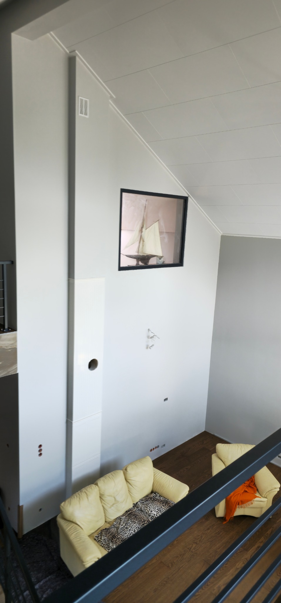 Wnętrze domu w trakcie wykańczania: białe ściany, drewniana podłoga, kremowe sofy, obraz z żaglówką, widok z góry zza barierki schodów.