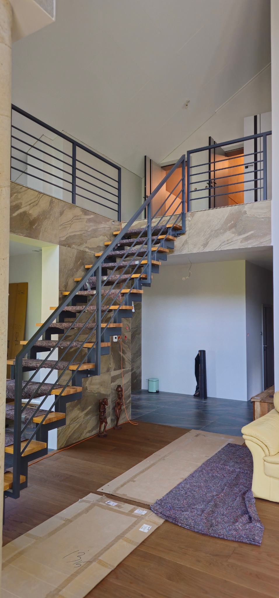 Dwupoziomowe wnętrze z nowoczesnymi schodami z metalową balustradą i drewnianymi stopniami z wykładziną, wykończone kamieniem dekoracyjnym, z widocznymi elementami zabezpieczającymi podłogę podczas...