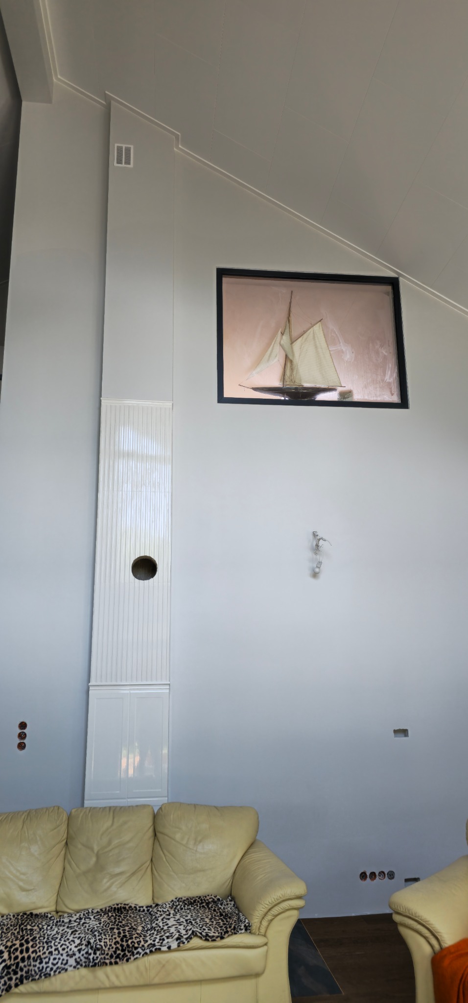 Wysoki salon z ukośnym sufitem, pomalowany na szaro, z dekoracyjną kolumną z otworem, obrazem żaglówki w czarnej ramie i kremową skórzaną sofą z narzutą w panterkę.