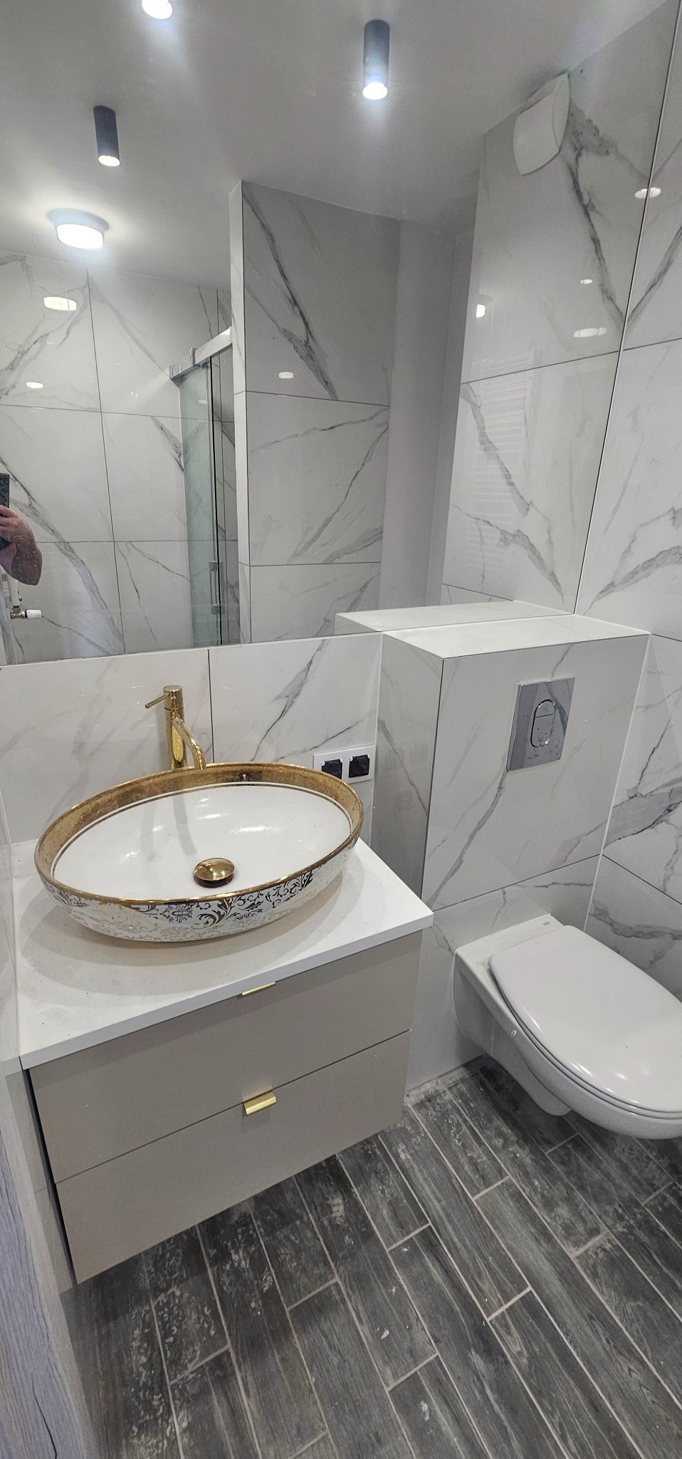 Elegancka łazienka po remoncie z owalną umywalką w kolorze białym ze złotymi zdobieniami, złotą baterią, podwieszaną toaletą i płytkami imitującymi marmur na ścianach.