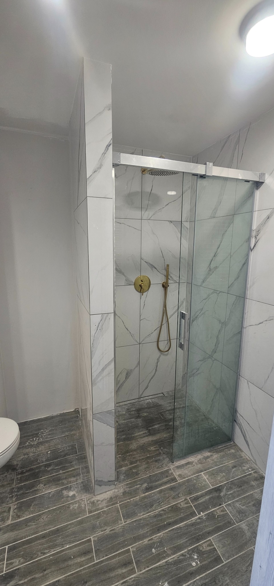 Nowoczesna kabina prysznicowa ze złotą armaturą i przesuwnymi drzwiami, wykończona płytkami imitującymi marmur na ścianach i płytkami imitującymi drewno na podłodze, widoczne ślady po remoncie.