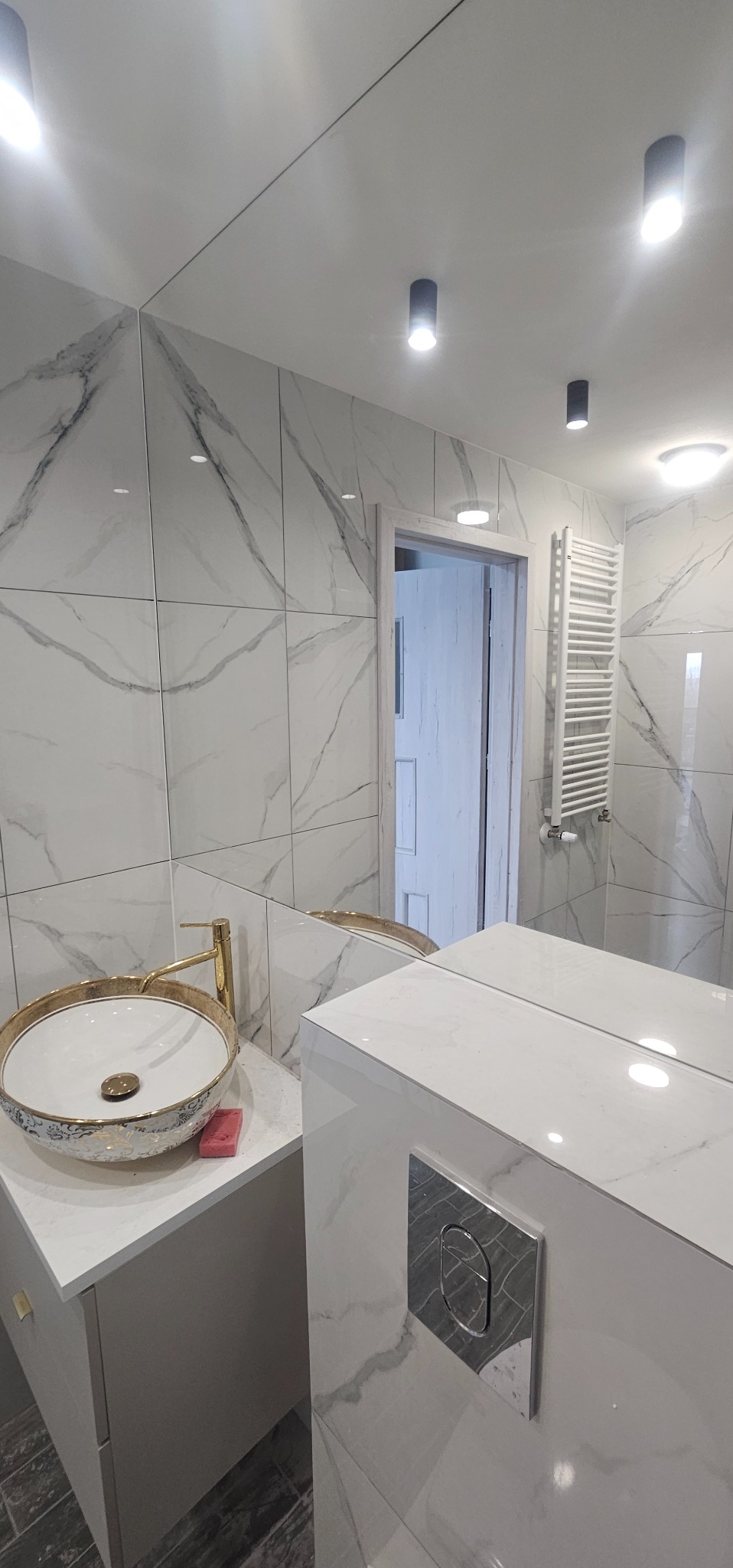 Nowoczesna łazienka z marmurowymi płytkami, ozdobną umywalką nablatową z elementami złota, chromowanym przyciskiem spłuczki, oraz podwieszanym grzejnikiem drabinkowym.