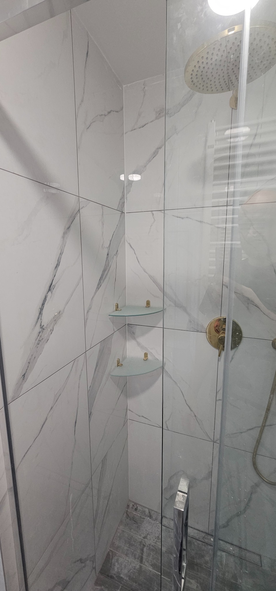 Szklana kabina prysznicowa z deszczownicą i półkami narożnymi w łazience wyłożonej białymi płytkami imitującymi marmur z szarymi żyłami, widoczny odpływ liniowy w podłodze.