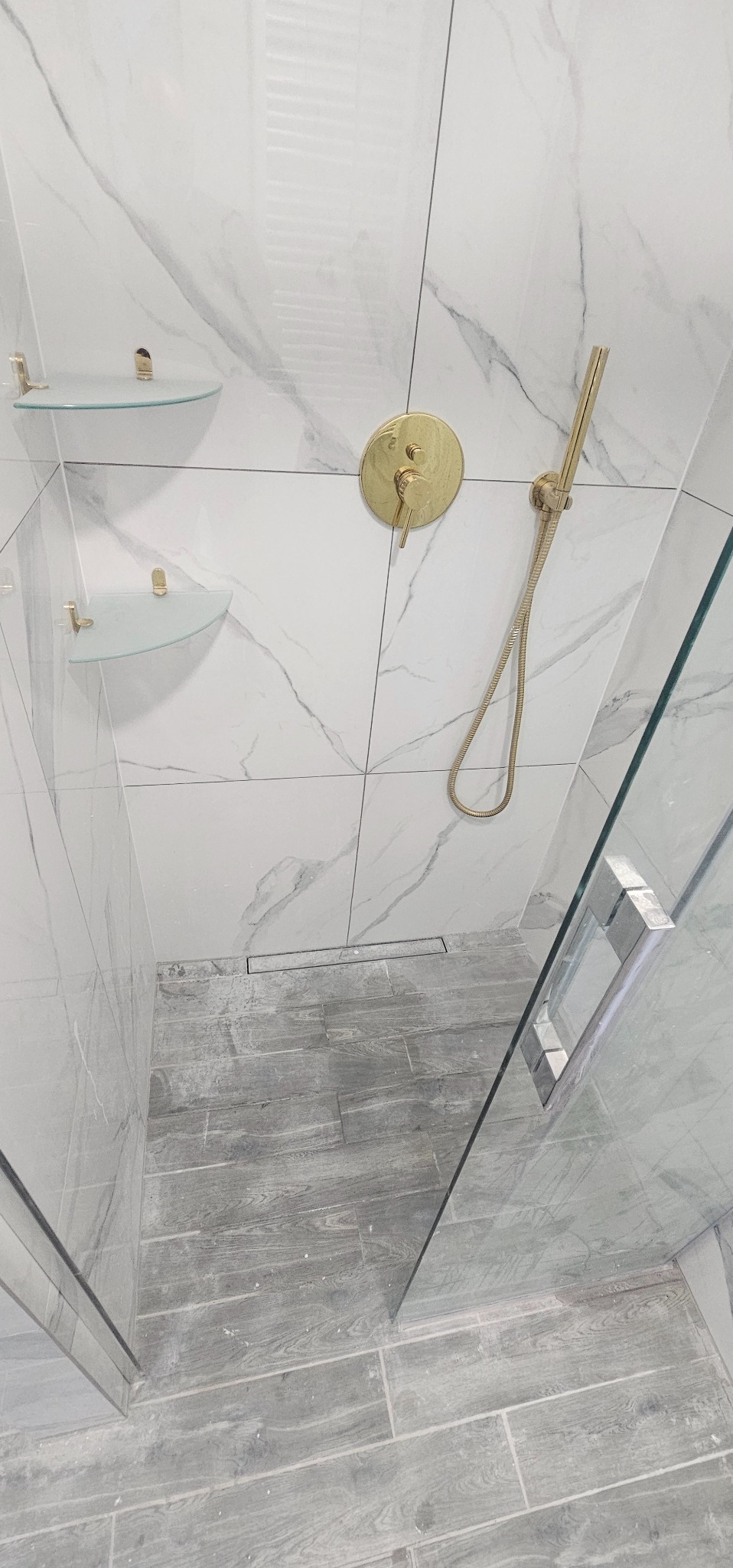 Nowoczesna kabina prysznicowa z białymi płytkami imitującymi marmur, złotą armaturą i szklanymi półkami narożnymi, wykończona szarymi płytkami podłogowymi imitującymi drewno.