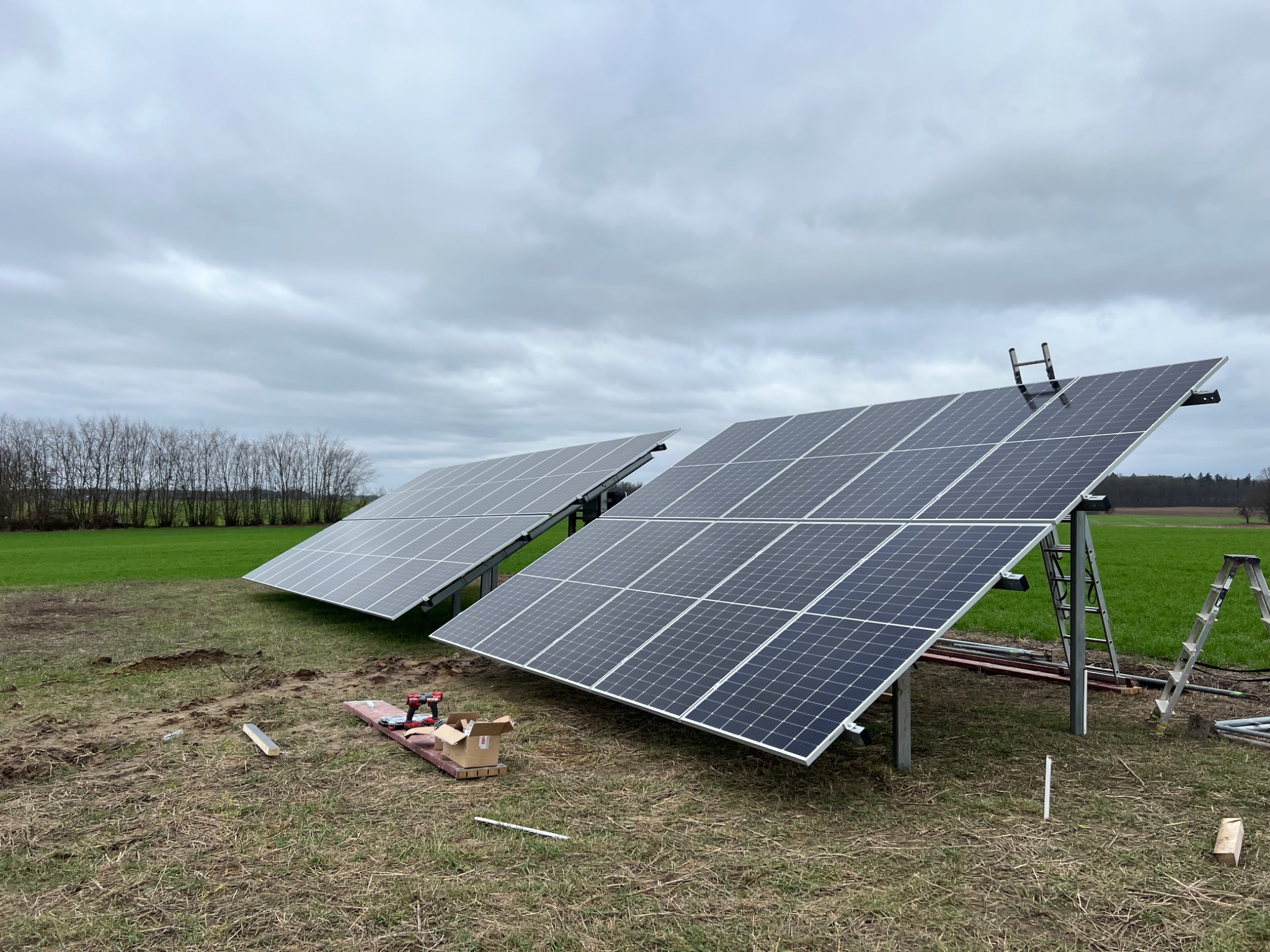 Dwa panele słoneczne na metalowych konstrukcjach, drabina oparta o jeden z paneli, narzędzia i karton na trawie, pochmurne niebo w tle.