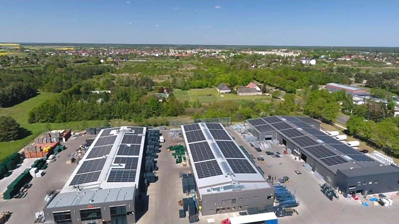 Widok z drona na budynki przemysłowe z panelami fotowoltaicznymi na dachach, z widocznym miastem i polami w tle.