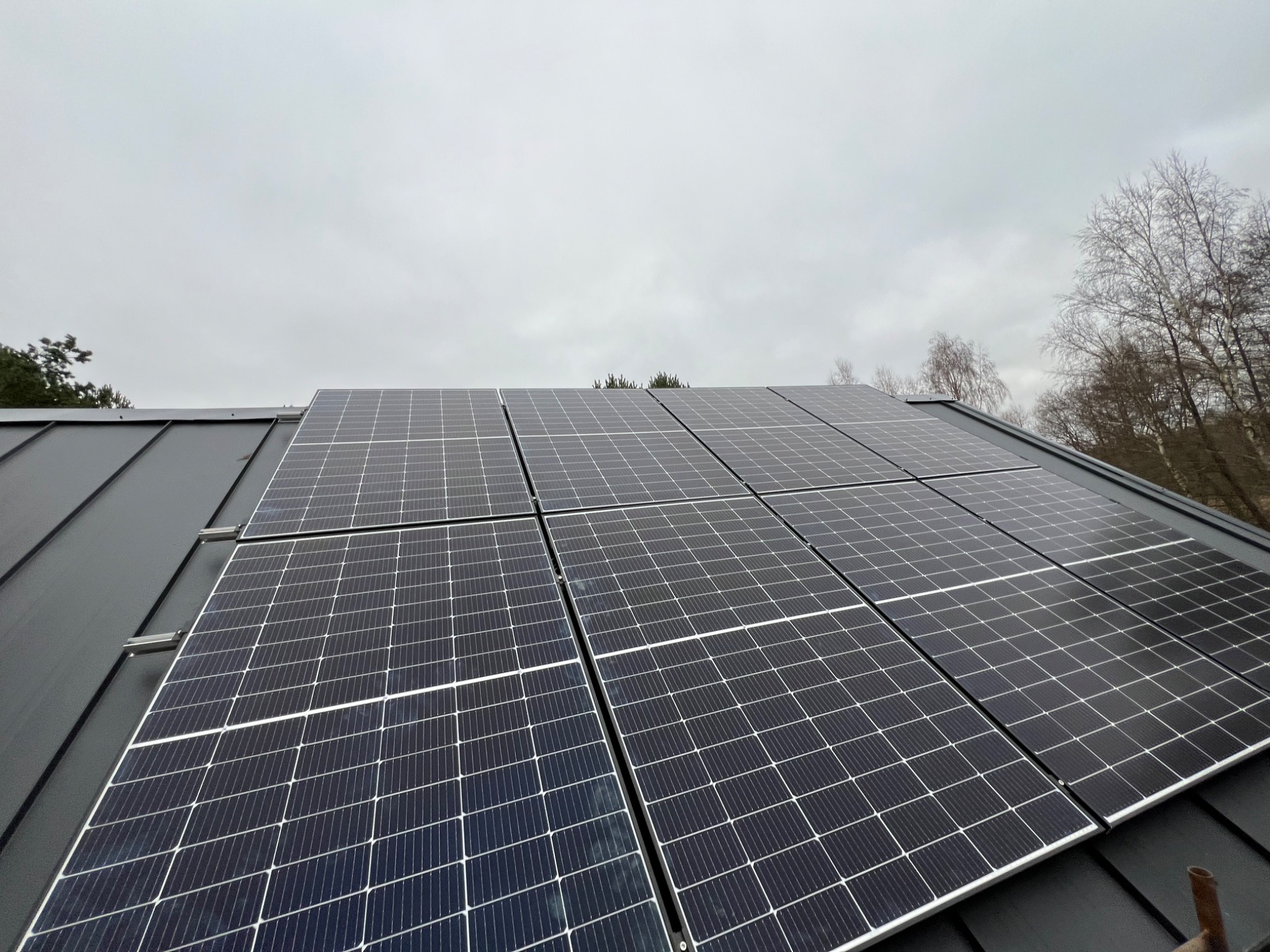 Czarny dach pokryty panelami słonecznymi w pochmurny dzień, widoczne drzewa w tle.