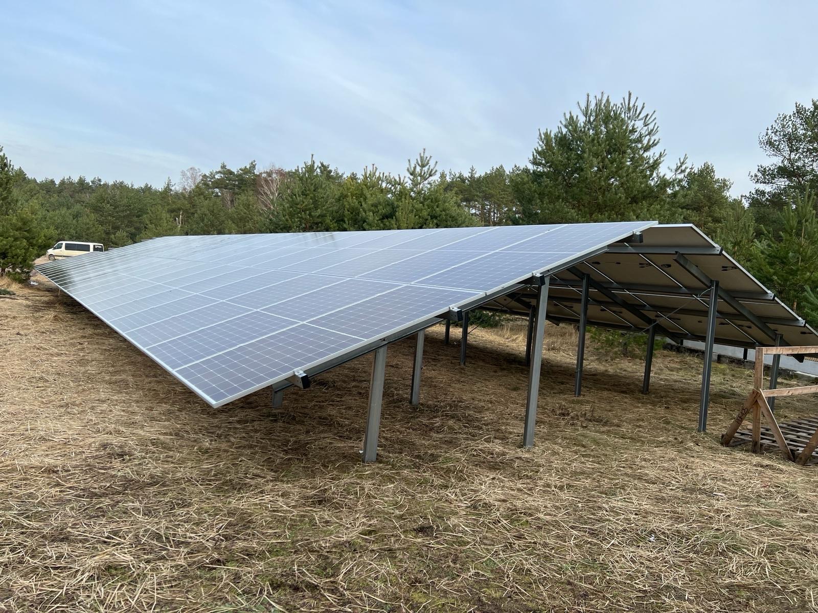 Rozległa instalacja paneli słonecznych na metalowej konstrukcji, ustawiona na nierównym terenie pokrytym suchą trawą, z lasem iglastym w tle i białym busem zaparkowanym w oddali.
