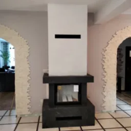 Nowoczesny, minimalistyczny wkład kominkowy z obudową z ciemnego kamienia, wkomponowany w jasne wnętrze z dekoracyjnymi łukami wykończonymi imitacją cegły.