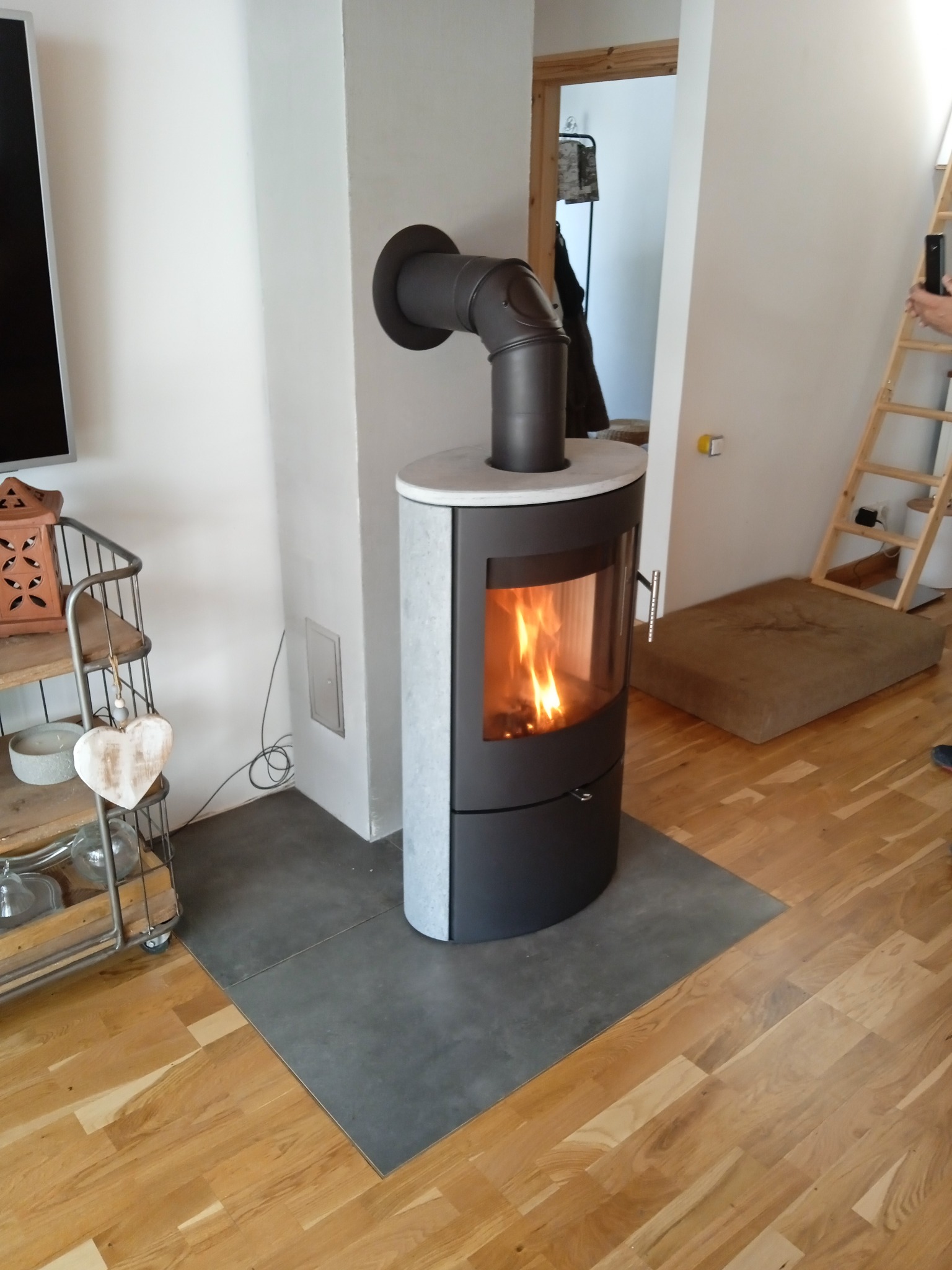 Nowoczesny piec wolnostojący z płomieniami wewnątrz, podłączony do komina, na tle jasnego wnętrza z drewnianą podłogą i drabinką.