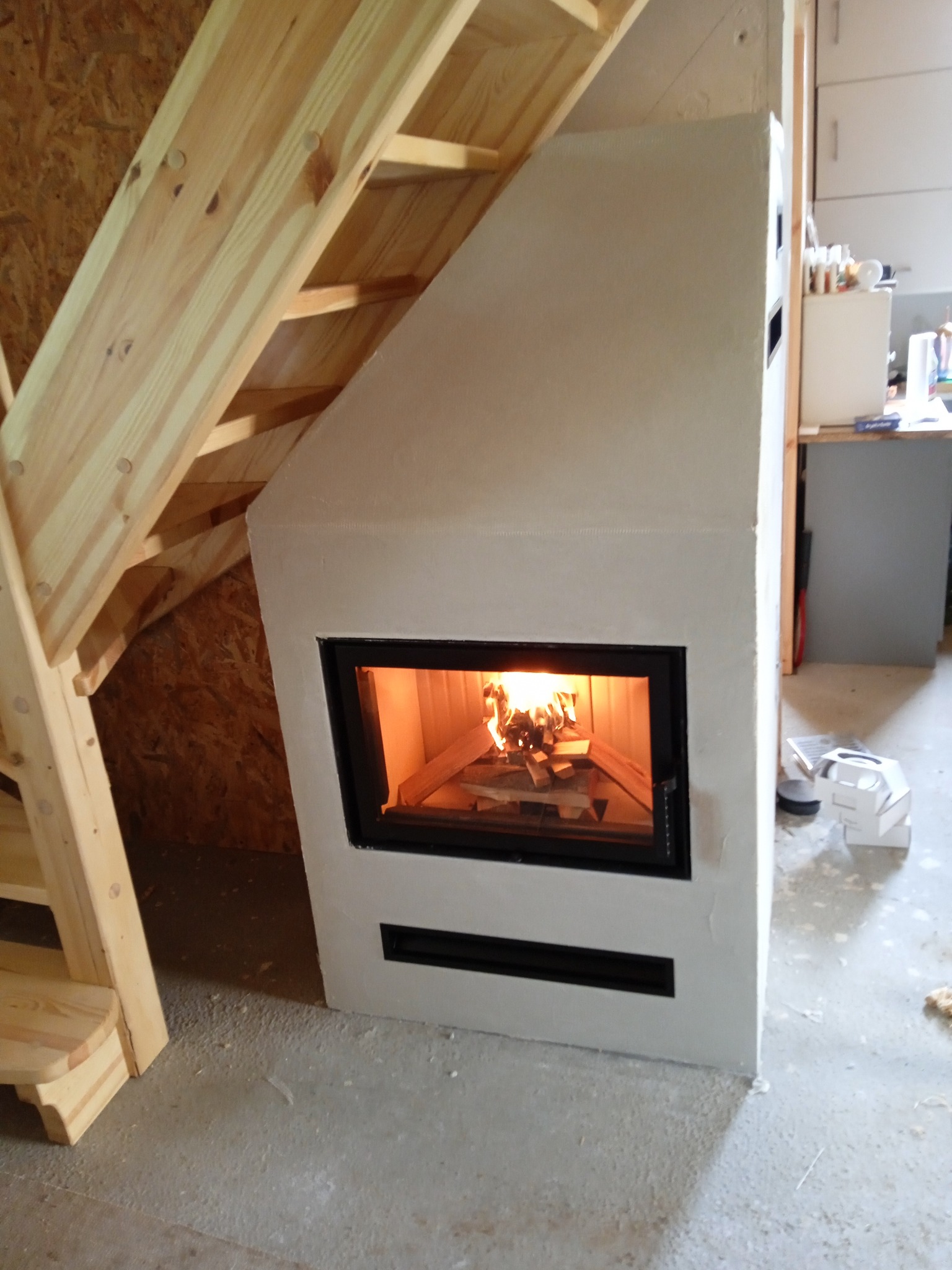 Biały, zabudowany piec z czarną ramą paleniska, płomienie wewnątrz, drewniane schody w tle, surowy beton na podłodze.