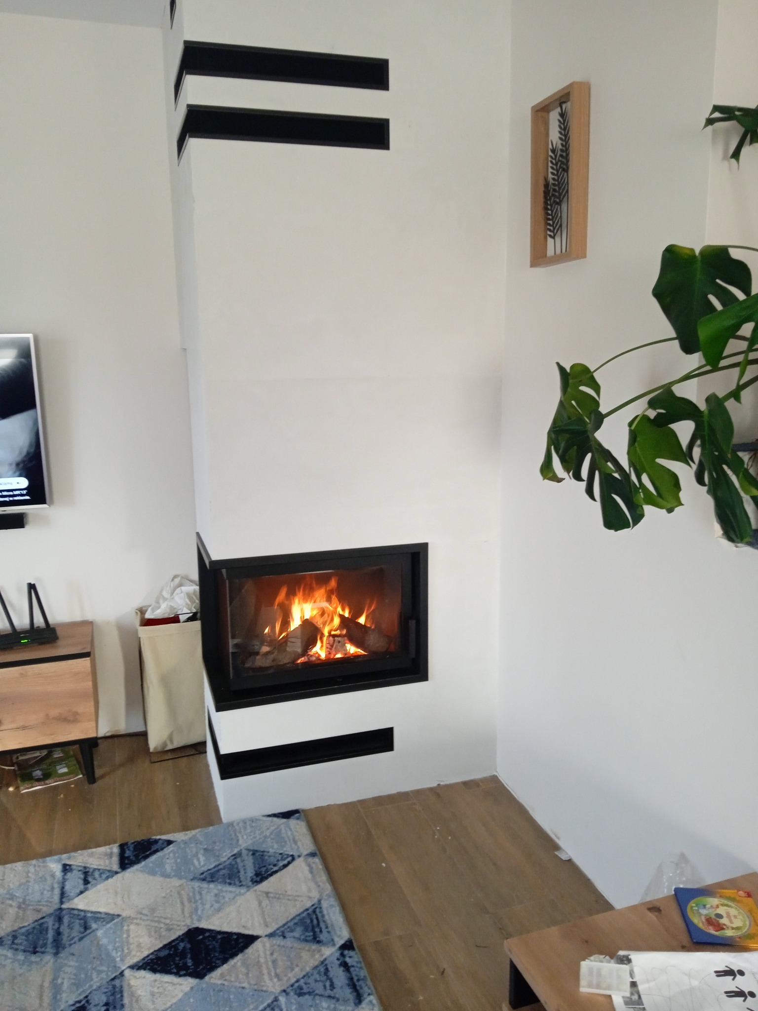 Wnętrze salonu z narożnym, nowoczesnym paleniskiem z czarną ramą i płomieniami, dekorowane poziomymi czarnymi pasami na białej obudowie, uzupełnione rośliną Monstera i obrazkiem na ścianie.