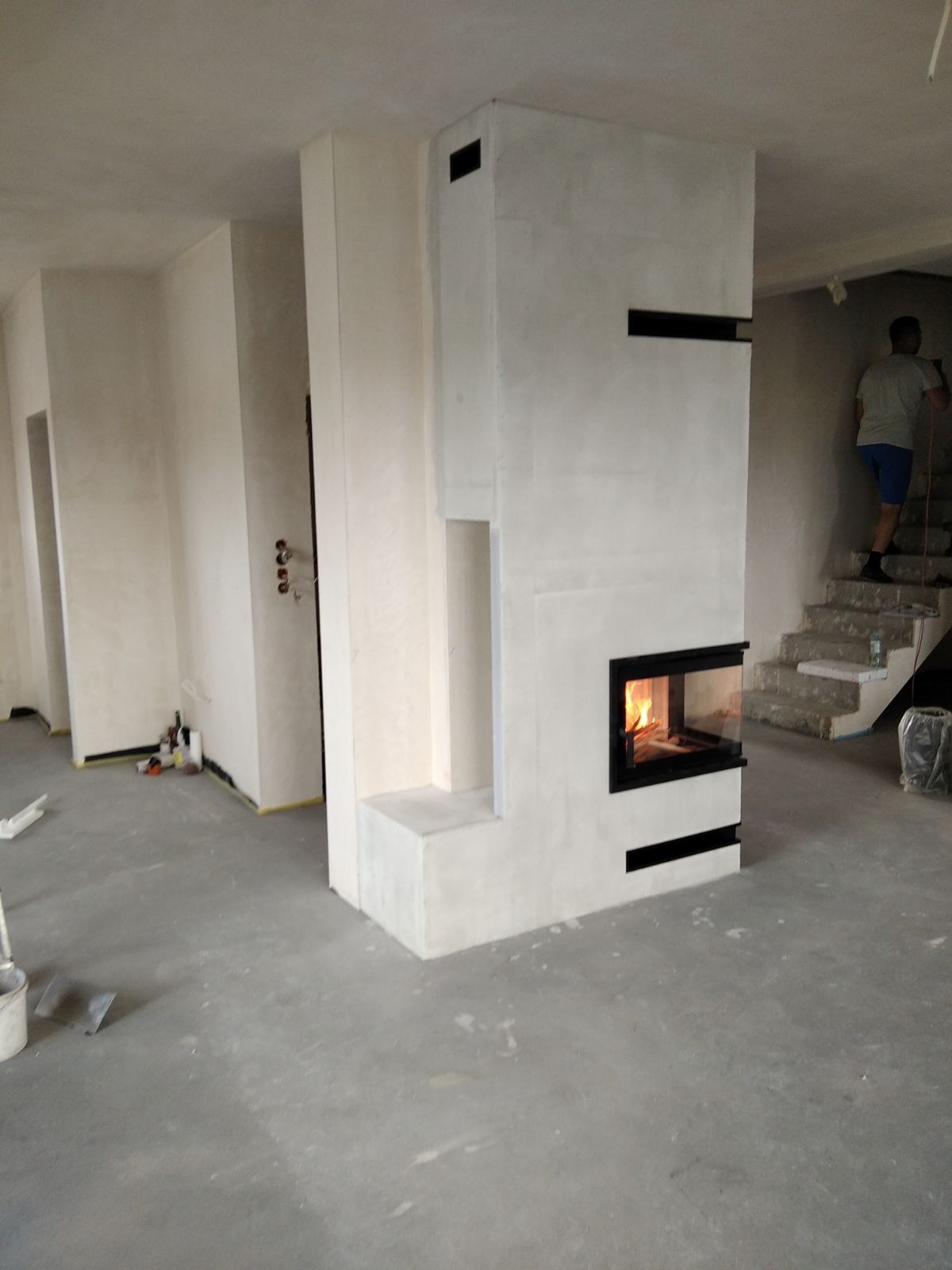 Nowoczesny, surowy w formie, biały piec z paleniskiem, w trakcie wykańczania wnętrza, widoczne schody i osoba w tle.