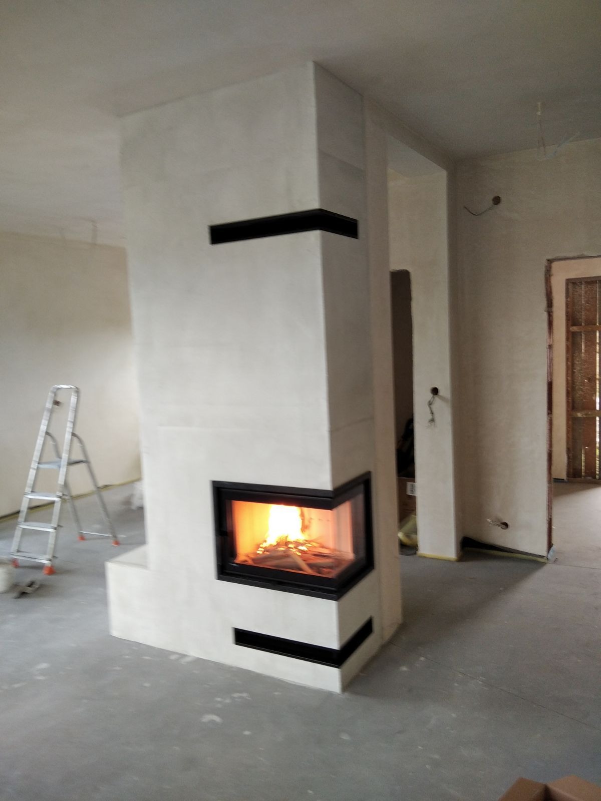 Nowoczesny, narożny wkład kominkowy z paleniskiem, obudowany jasnym kamieniem z czarnymi akcentami, w surowym wnętrzu z drabiną malarską w tle.
